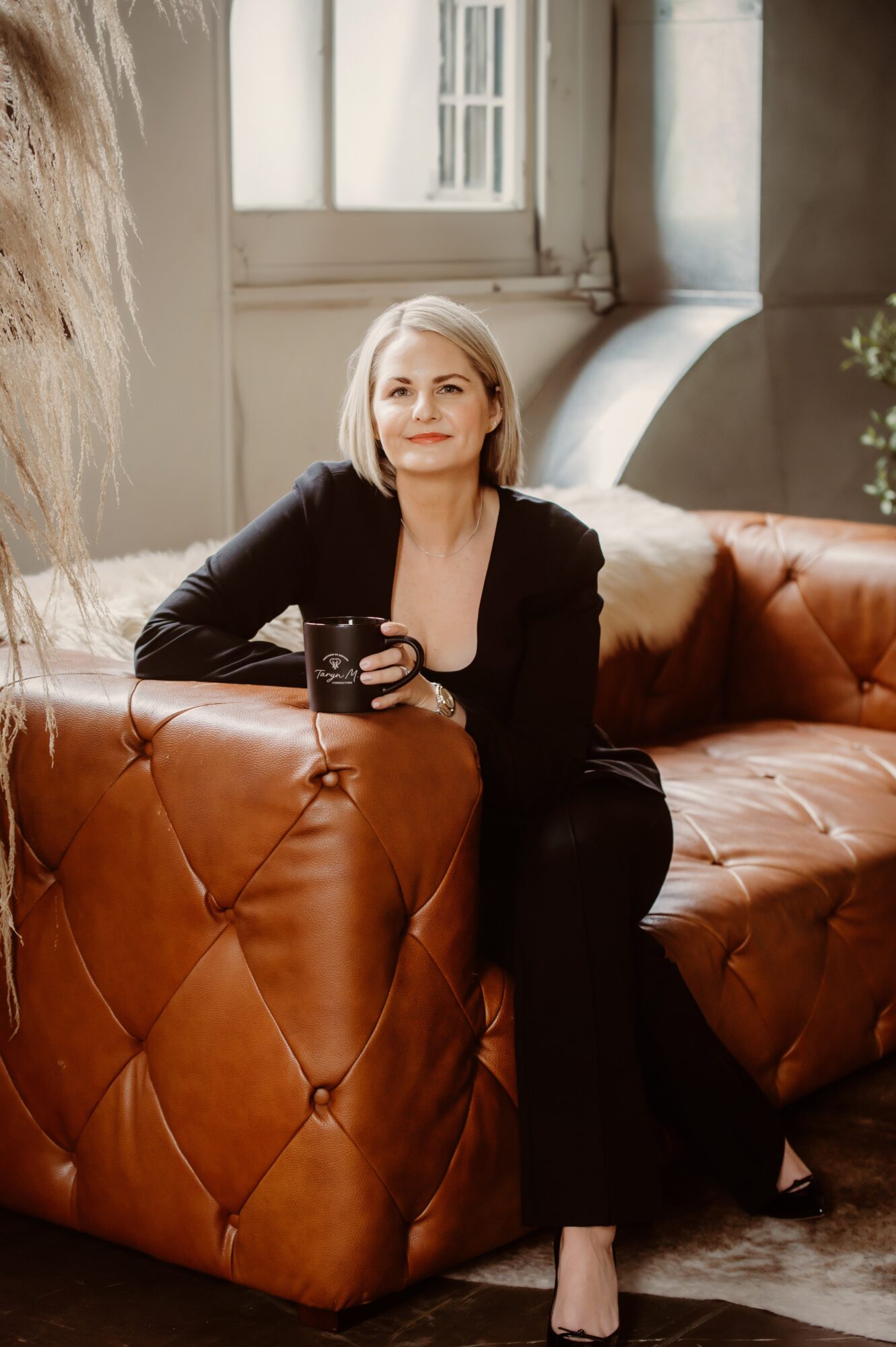 Woman with short blonde hair sitting on a brown leather sofa, holding a mug, in a bright room with large windows.