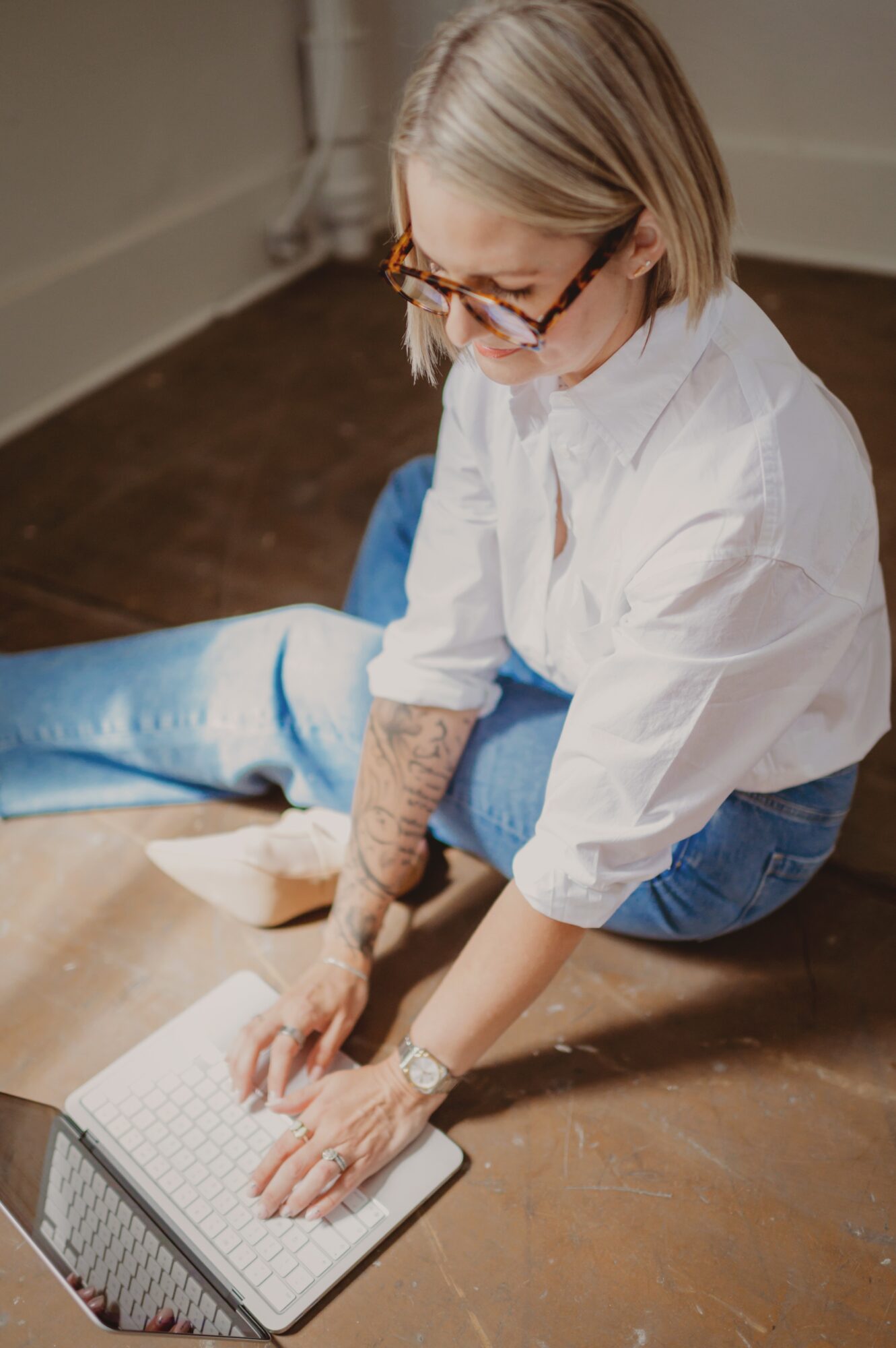 Person with glasses and tattoos on hands sitting on floor, using a laptop, wearing a white shirt and jeans.