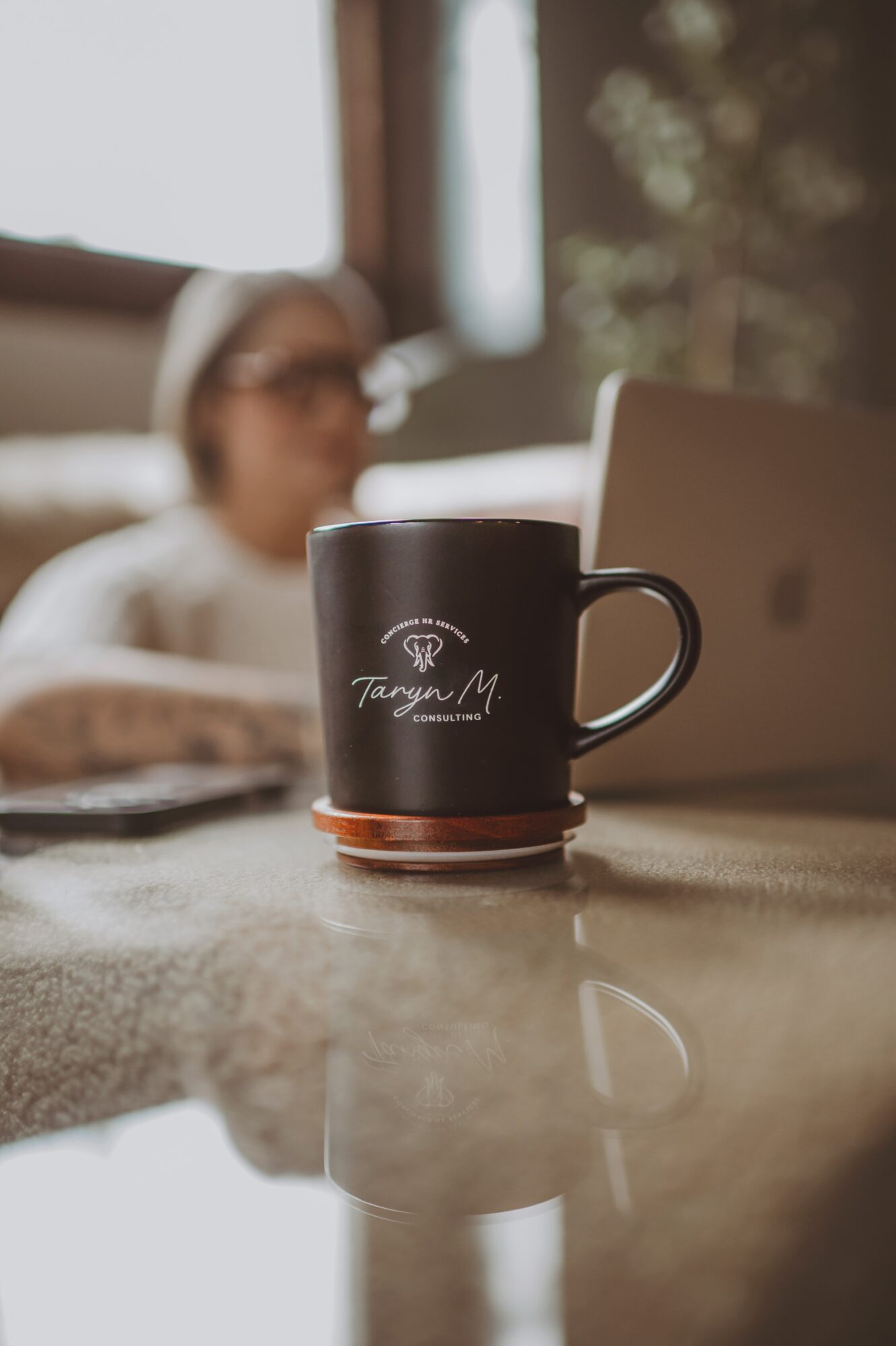 Black coffee mug with white text on a table, blurred person in background, laptop nearby, reflection on table surface.