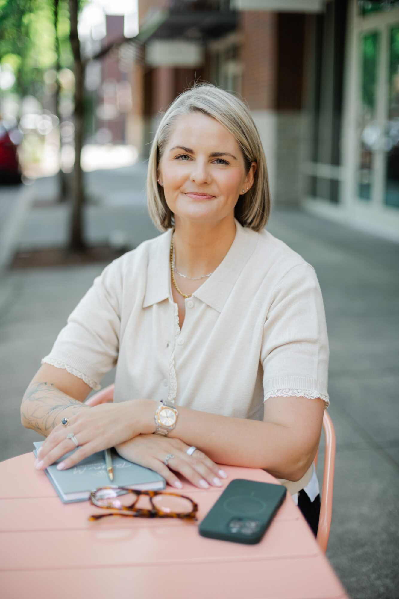 Woman with short blonde hair sitting at a table outdoors, smiling, with a smartphone and glasses on the table.
