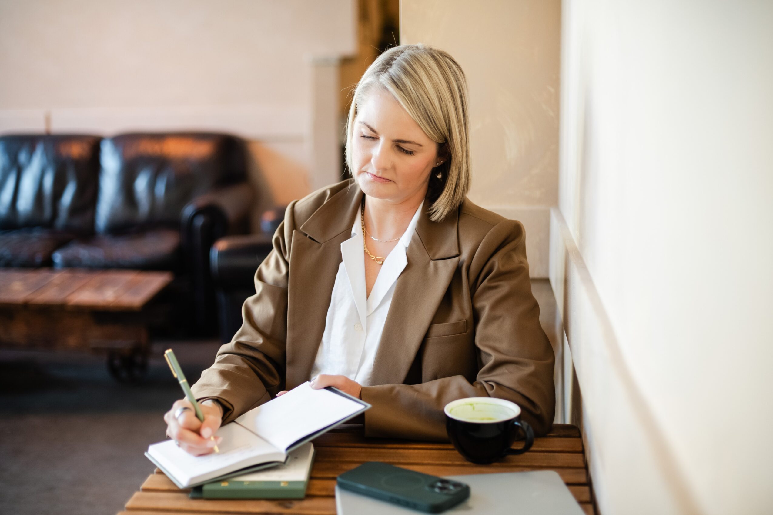 Woman with blonde hair in a brown blazer writing in a notebook at a wooden table, with a bowl and a phone nearby, in a cozy room.