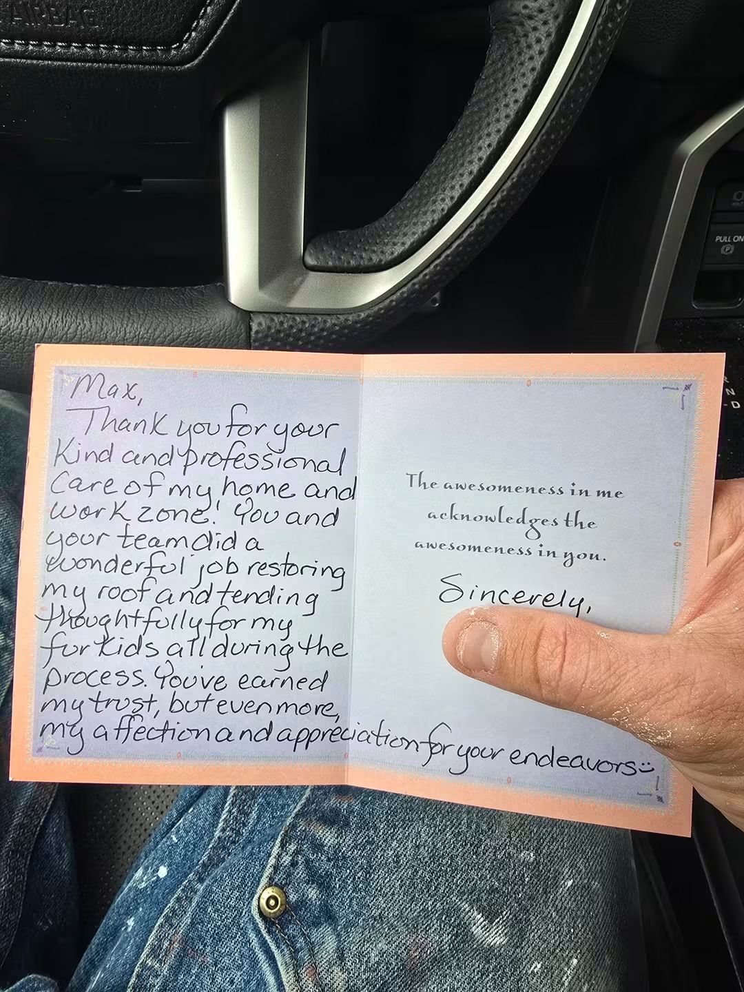 Handwritten note on pink paper inside a car, with a steering wheel and dashboard visible in the background.