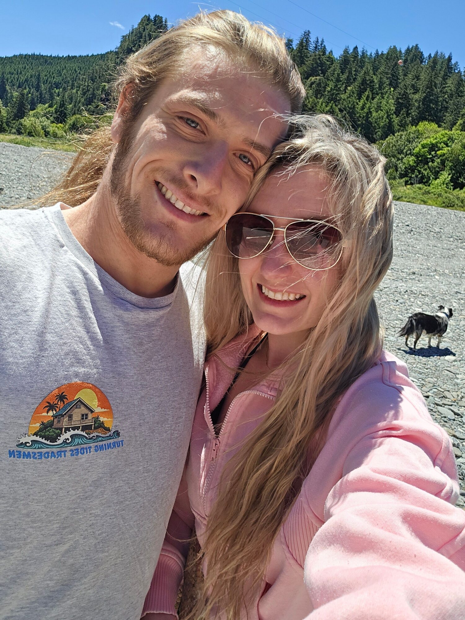 Two people smiling outdoors, one wearing sunglasses and a pink jacket, the other in a gray shirt with a logo, with trees and sky in background.