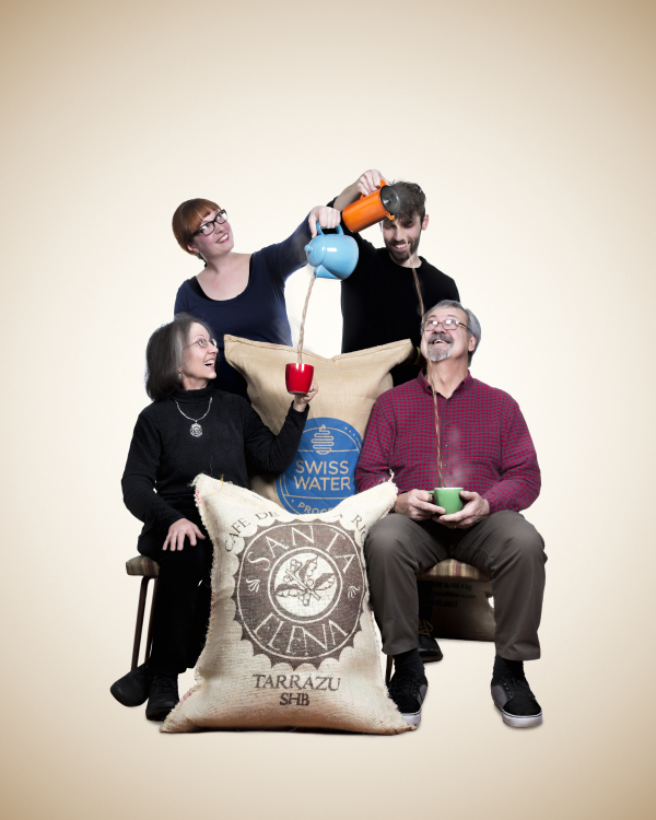 Four people with glasses and beards, holding cups and a water bottle, sitting and standing around a large bag of Swiss water, against a plain background.