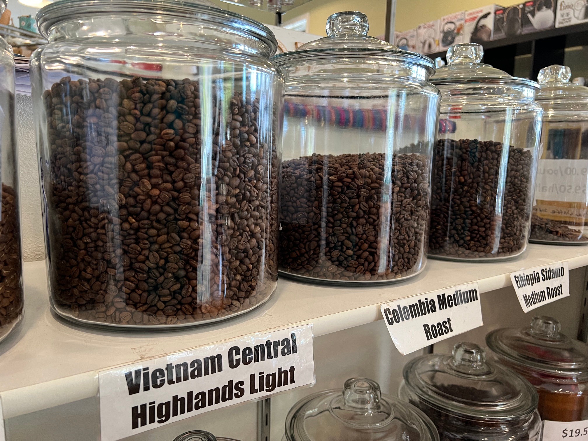 Glass jars filled with coffee beans on a shelf, labeled with different coffee roast names, in a store setting.