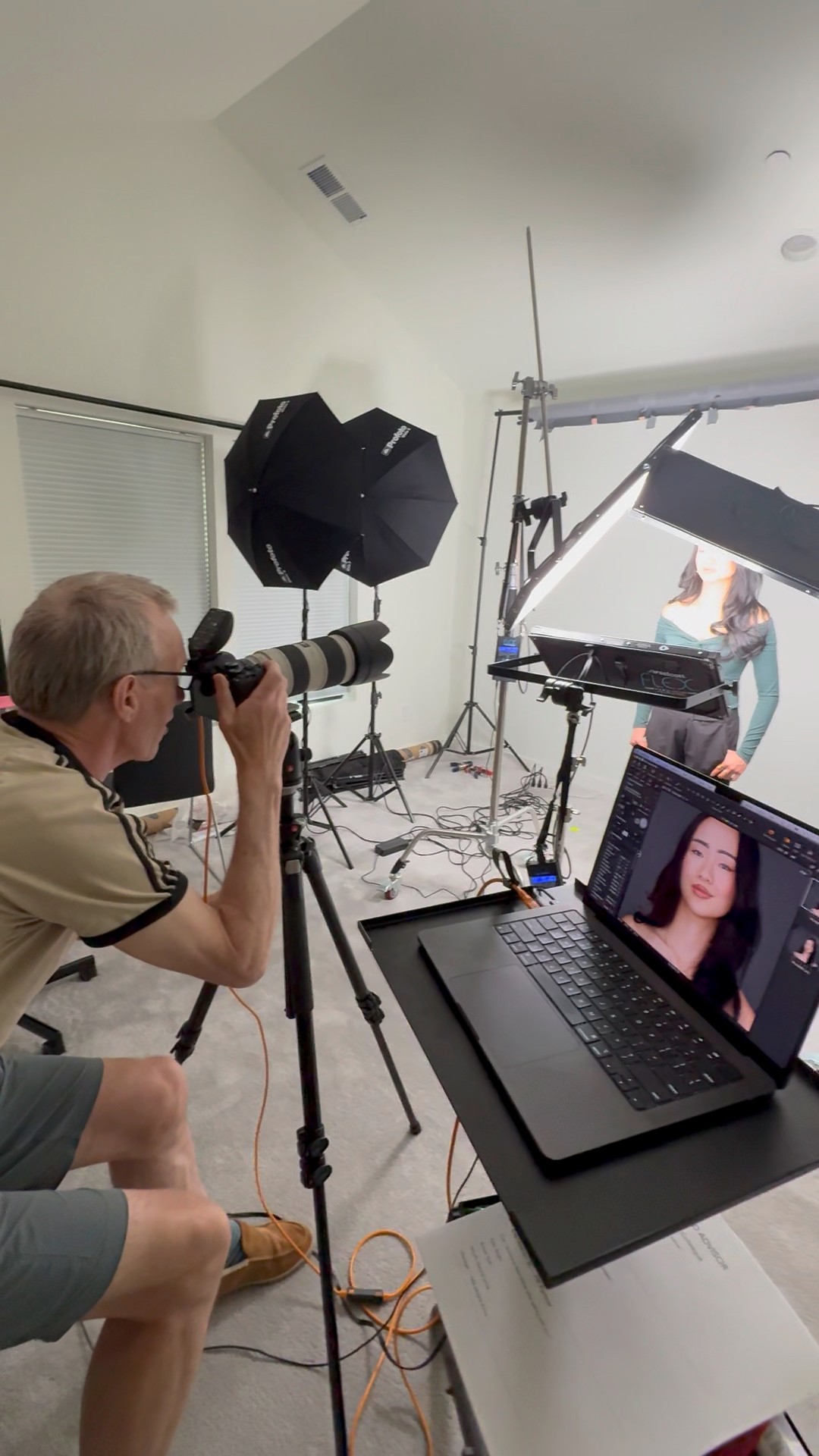 Photographer takes picture of a woman in a studio with lighting and equipment, laptop shows her face, studio background visible.