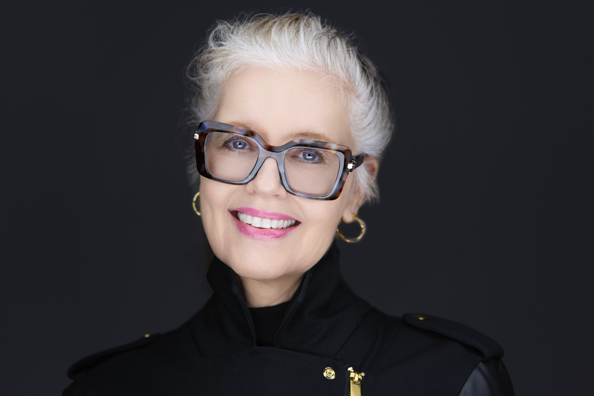 Portrait of an older woman with white hair, glasses, and gold earrings, smiling against a dark background.