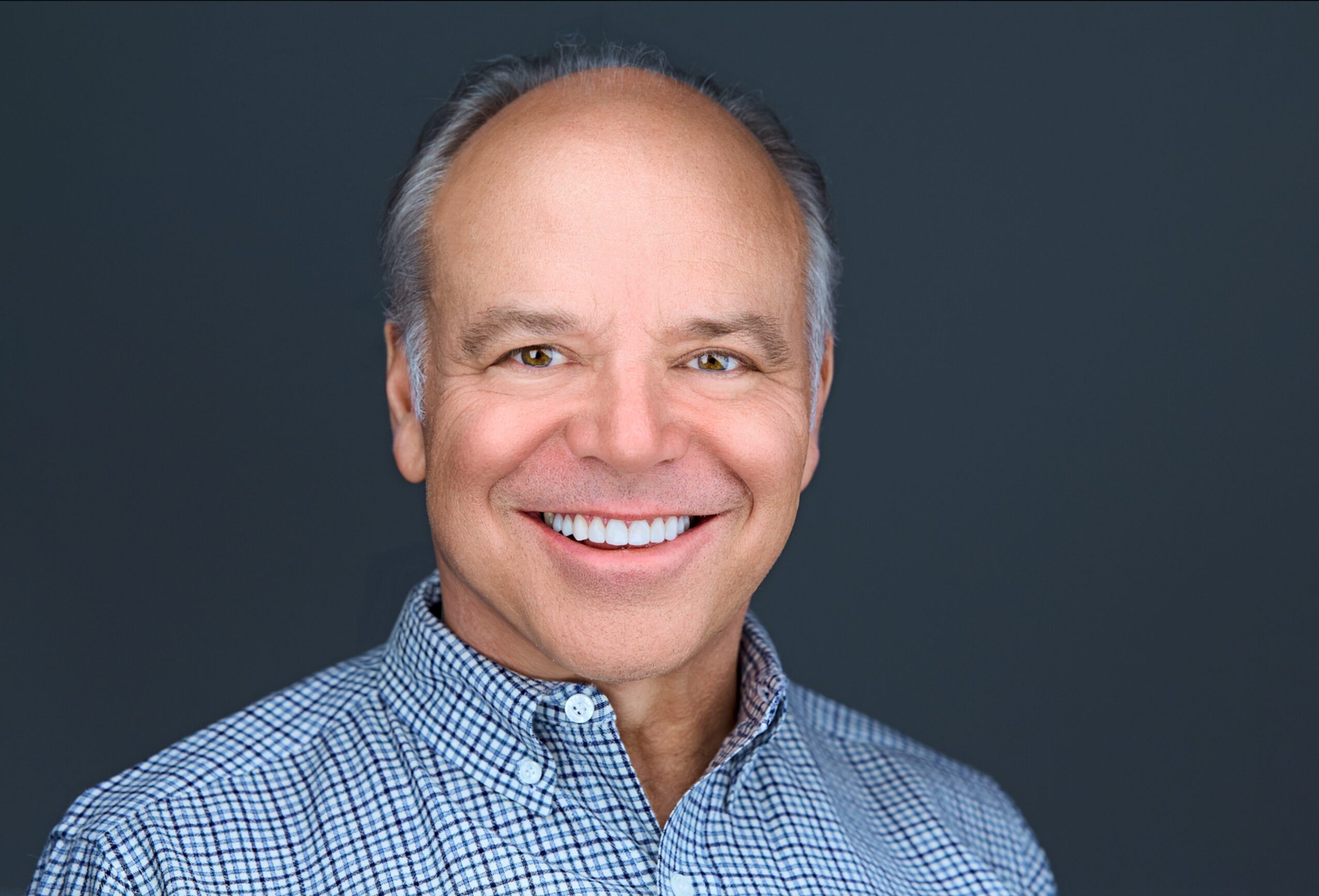 Smiling middle-aged man with short gray hair wearing a checkered shirt against a dark background.