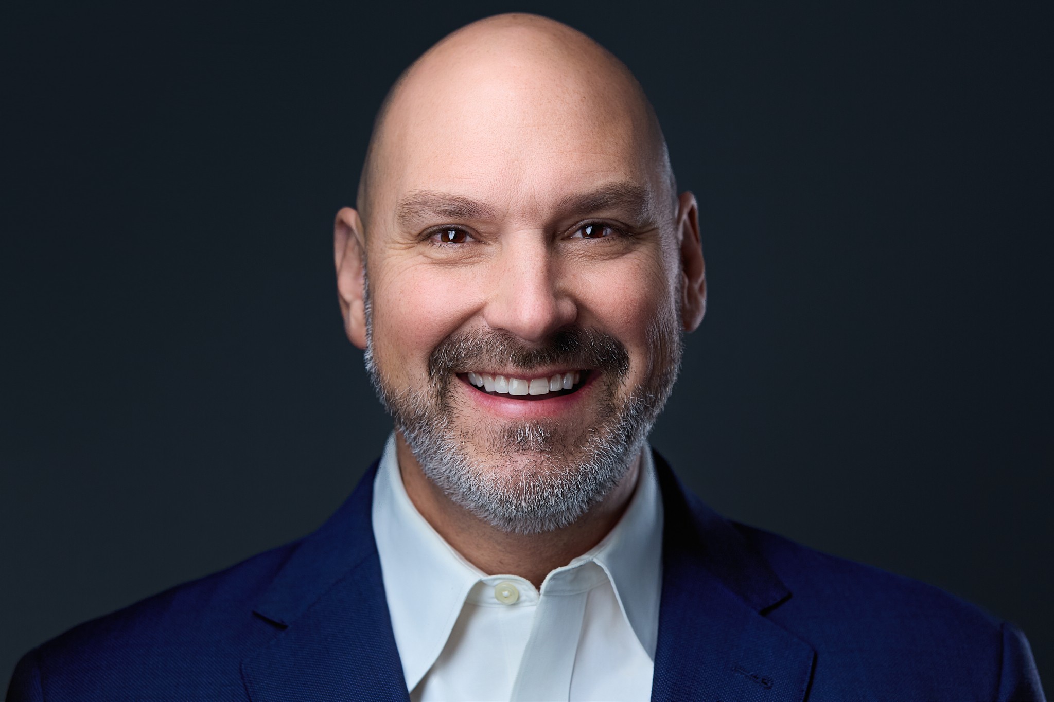 A smiling bald man with a beard wearing a dark suit and white shirt, against a dark background.