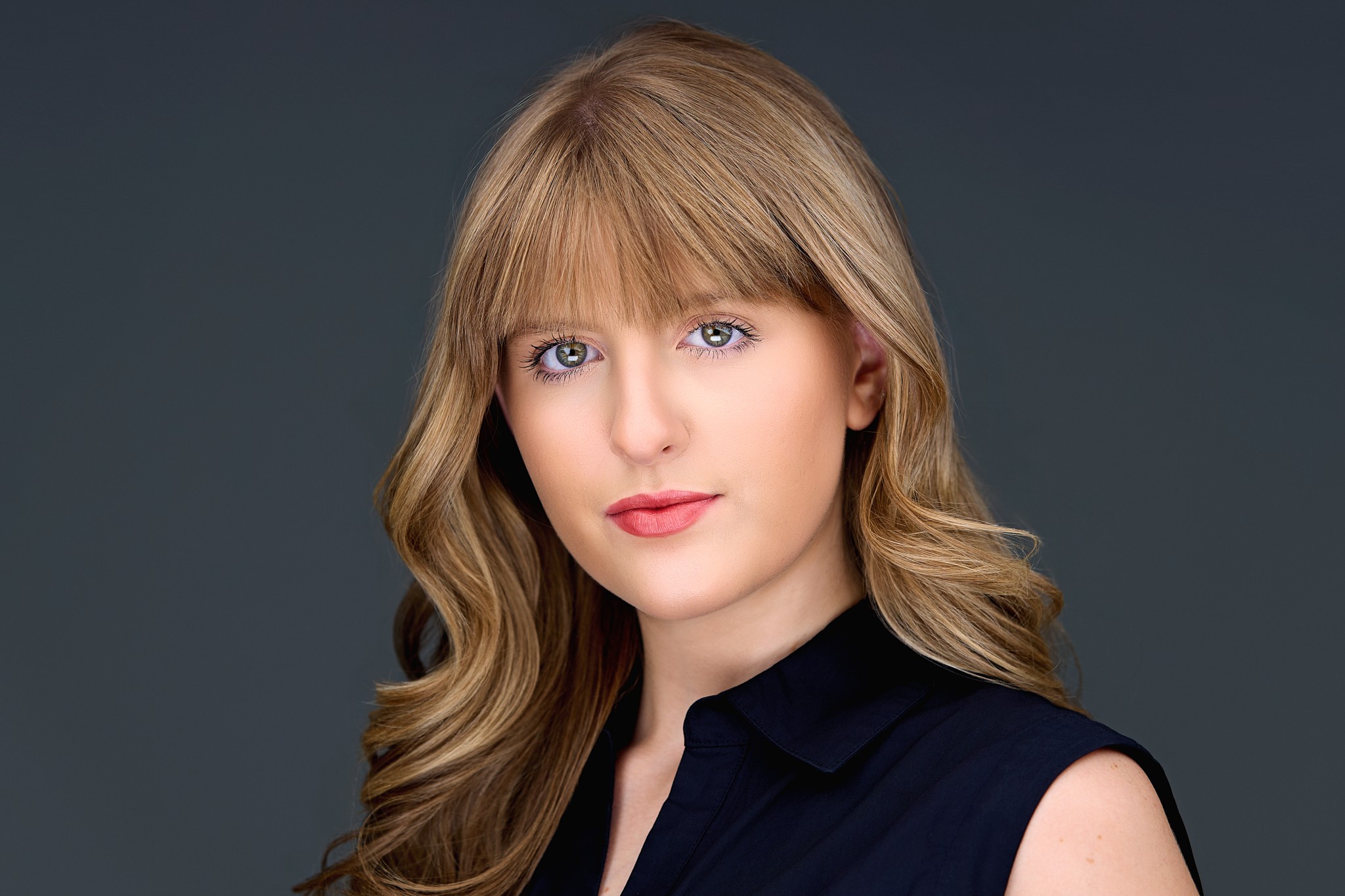 Young woman with blonde hair and blue eyes wearing a black sleeveless top, looking at the camera against a dark background.