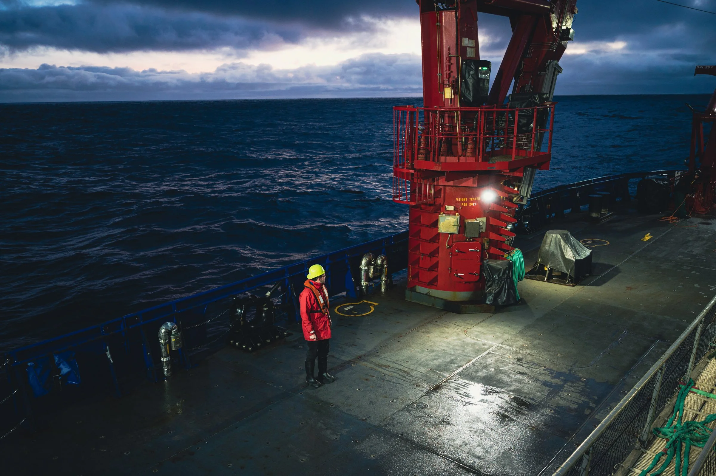 Person in safety gear stands on a ship's deck near a large red crane, with ocean and cloudy sky in background.