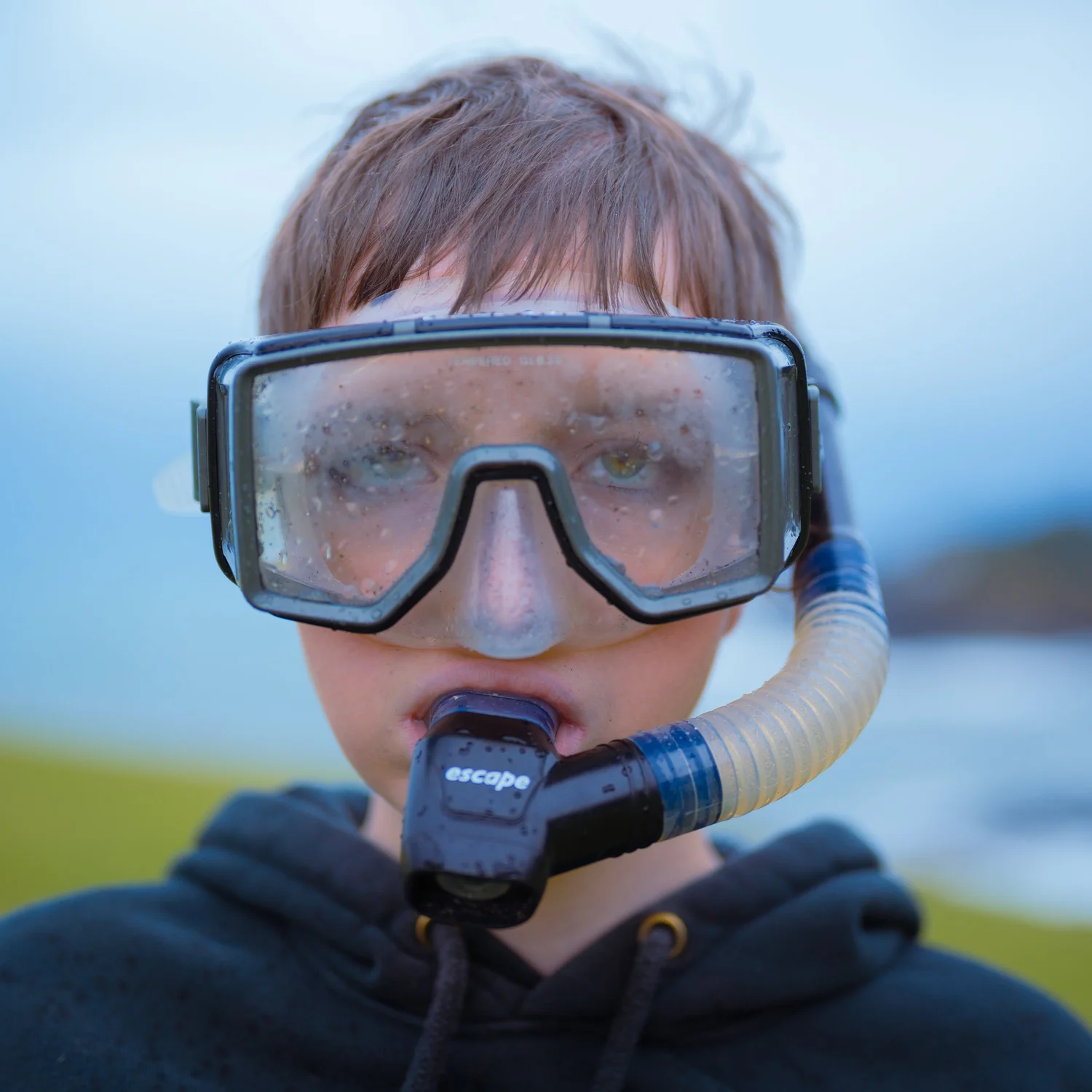 Child wearing a diving mask and snorkel outdoors, with a blurred background of sky and landscape.