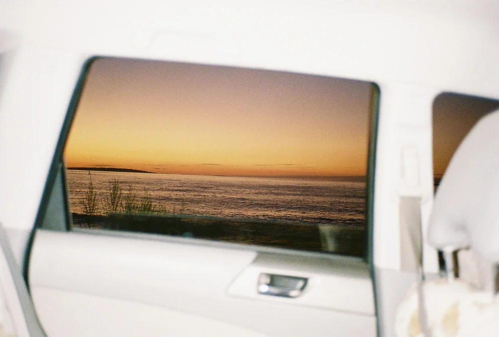 Sunset over a body of water with a grassy shoreline, viewed through a car window.