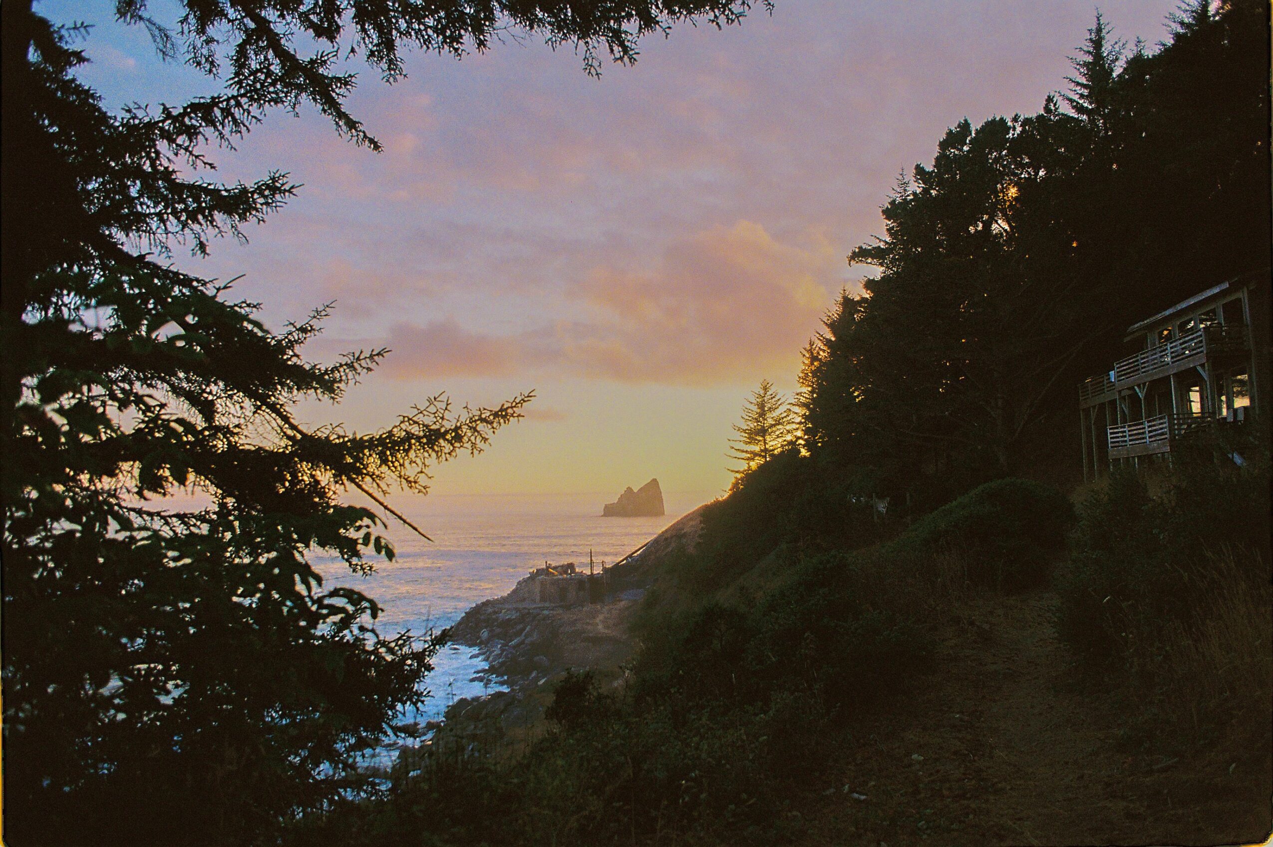 Sunset over ocean with rocky coastline, trees, and a small building on the hillside, sky with pink and purple hues.