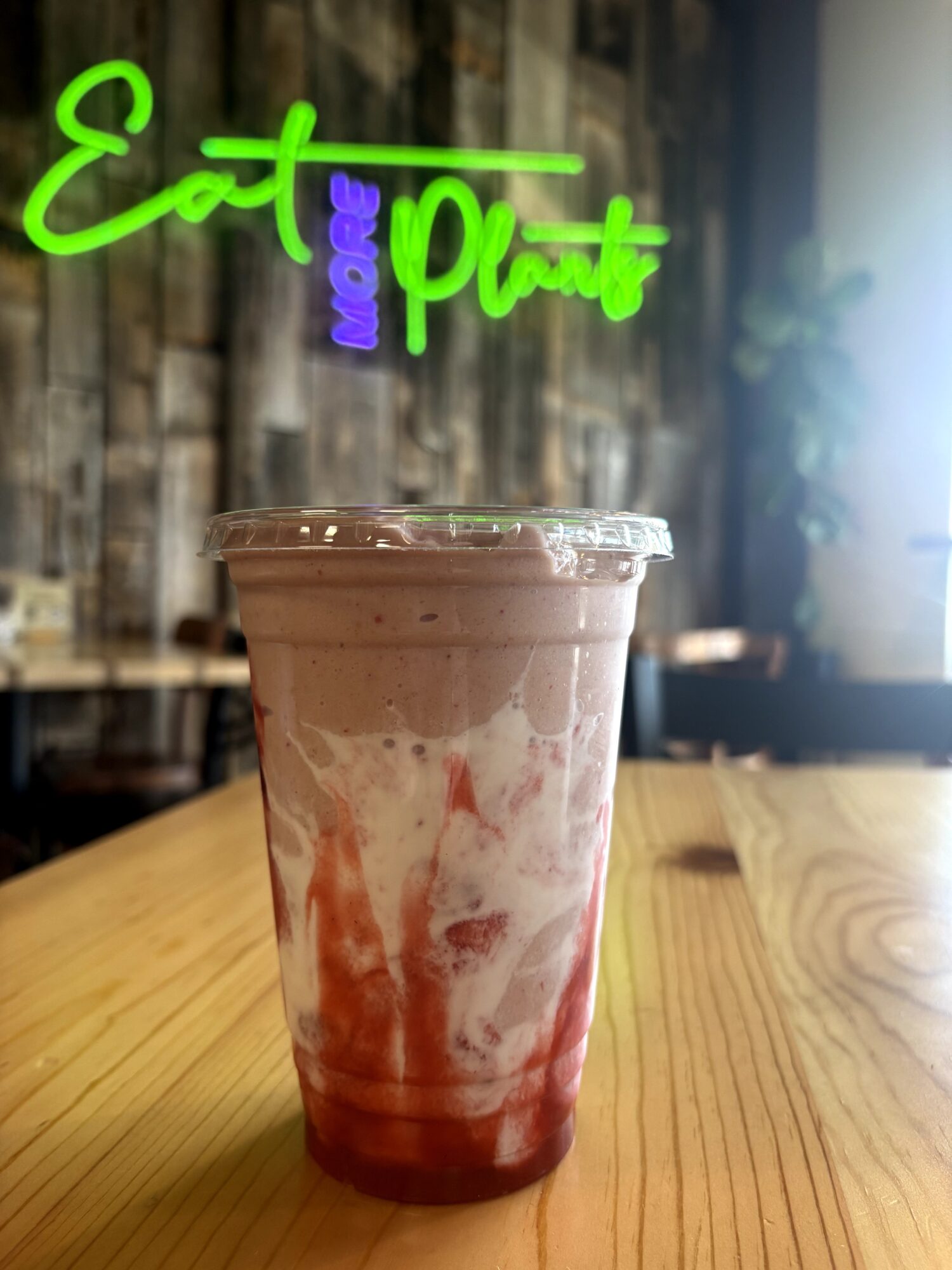 A plastic cup with a layered pink and white beverage on a wooden table, neon sign in background, interior setting.