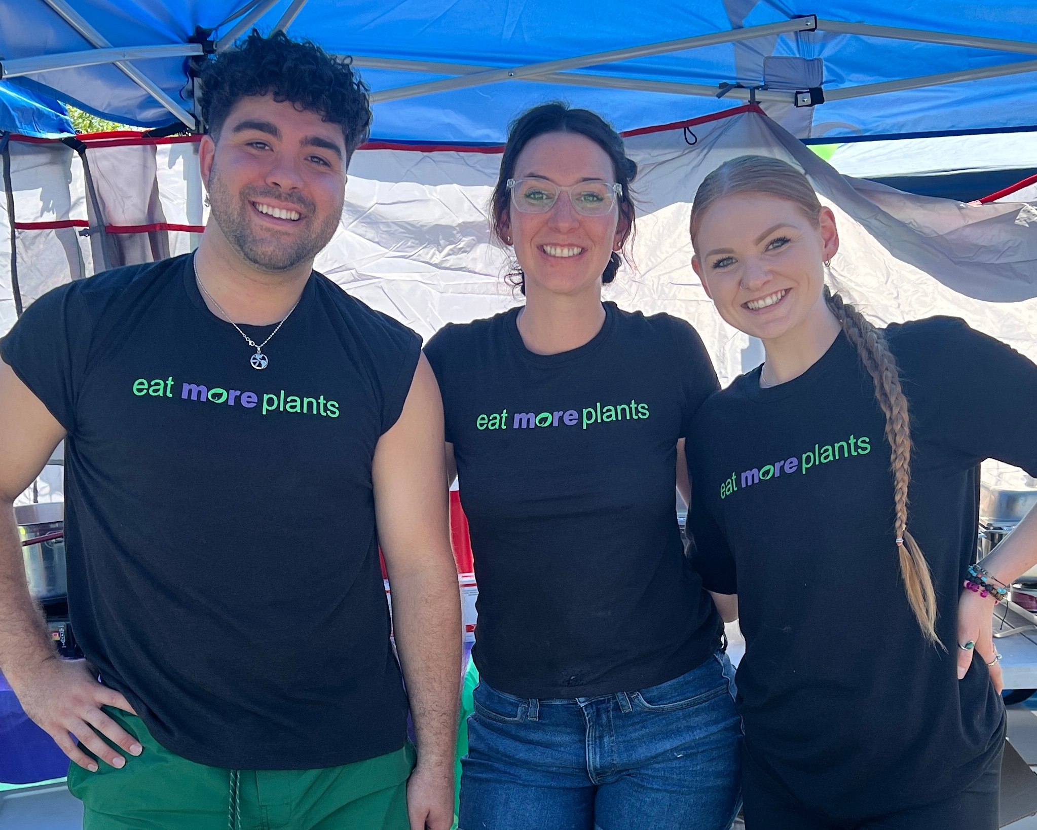 Three smiling people wearing black shirts with green and purple text, standing under a blue canopy.
