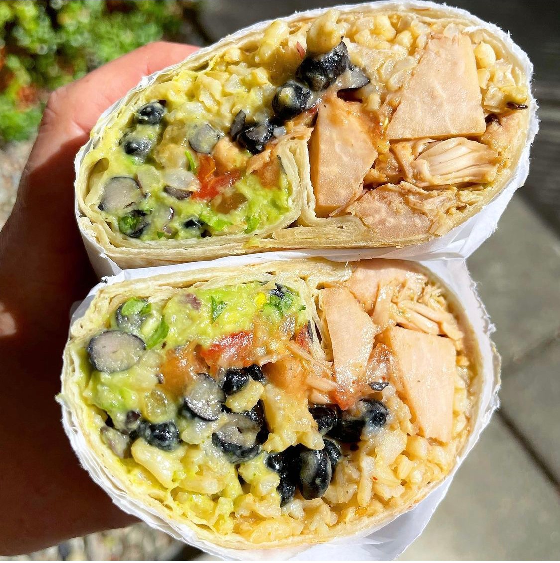 Two halves of a burrito with rice, black beans, chicken, and vegetables, held in a hand outdoors.