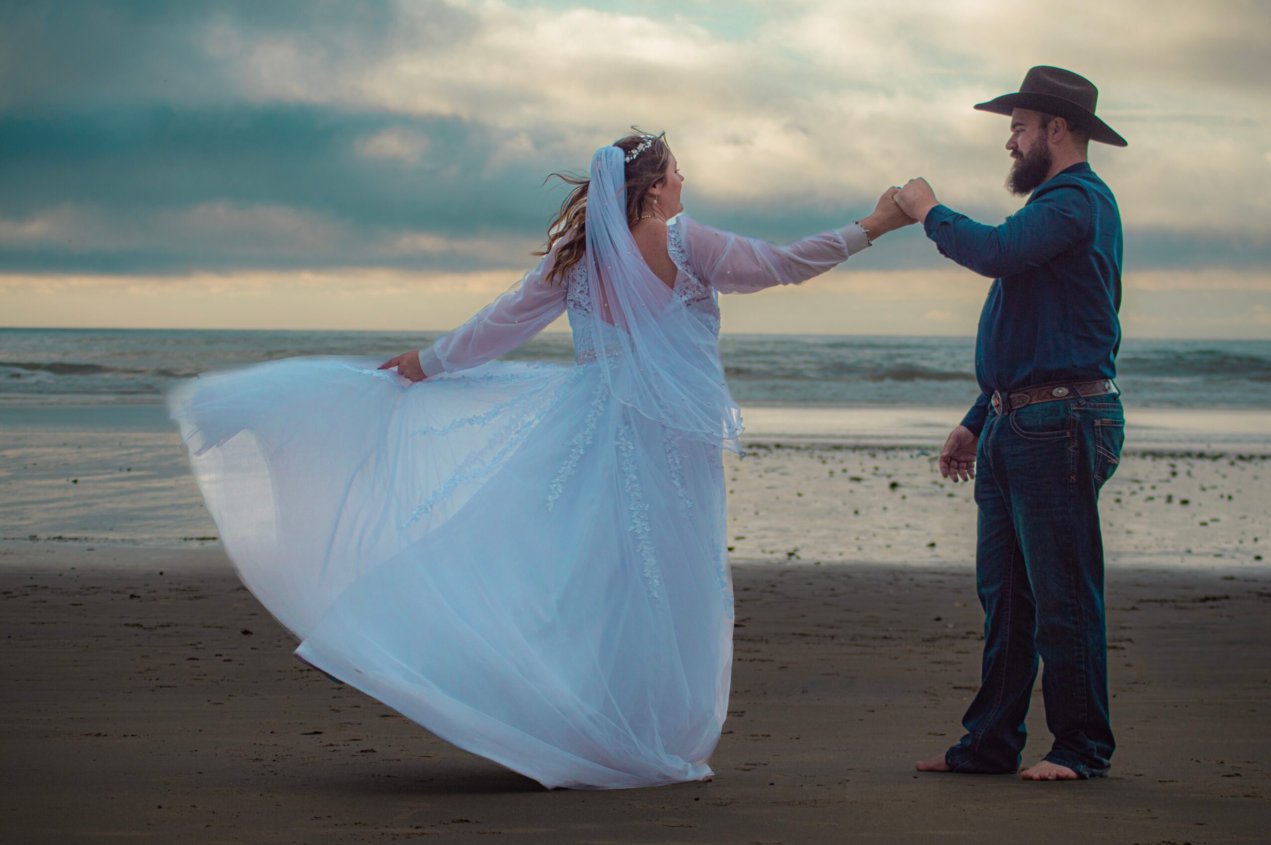 A couple dancing on the beach at sunset, woman in a flowing white dress and man in a cowboy hat and jeans, holding hands.