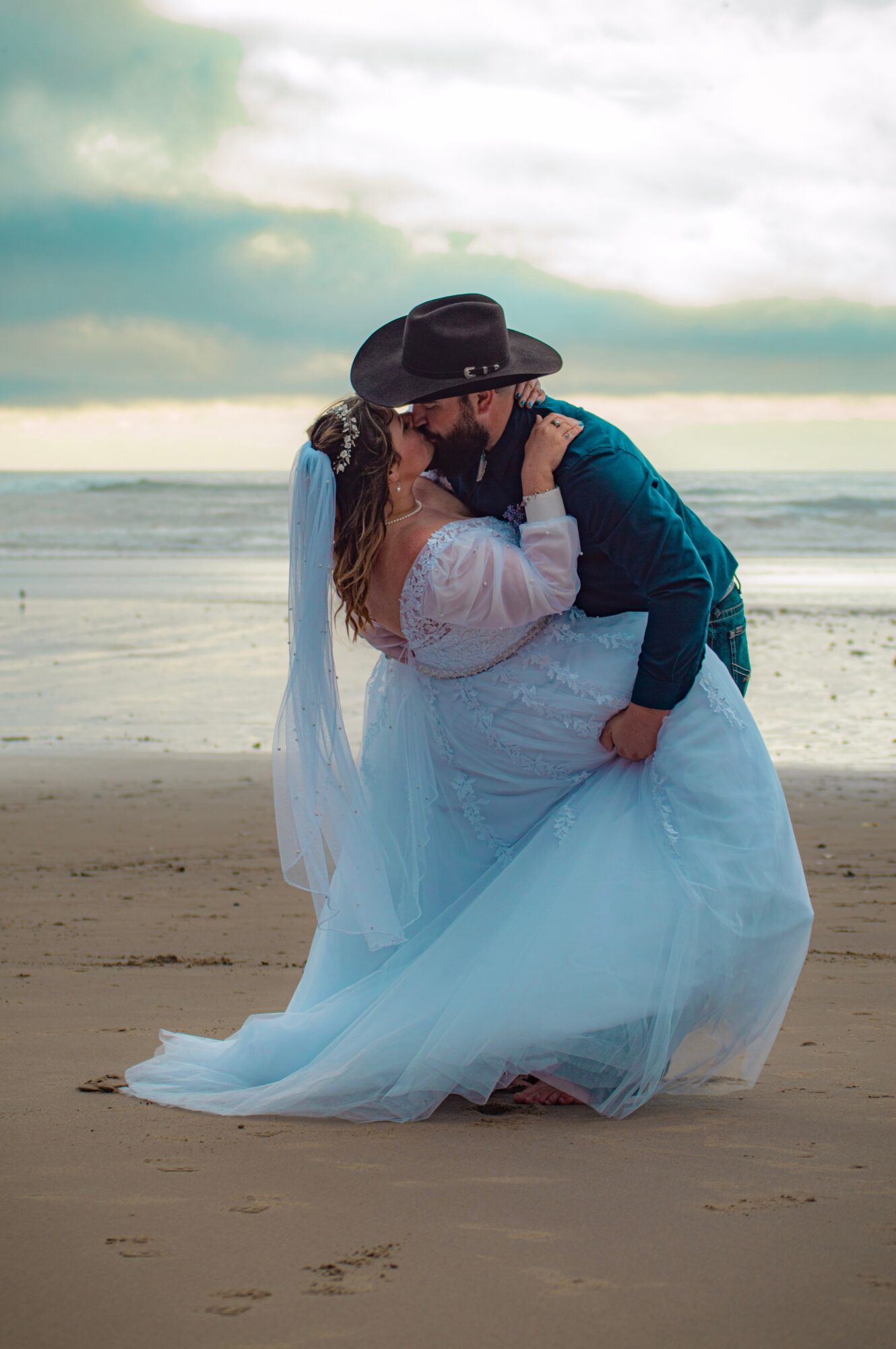 A couple in wedding attire kissing on the beach with ocean and sky in background.