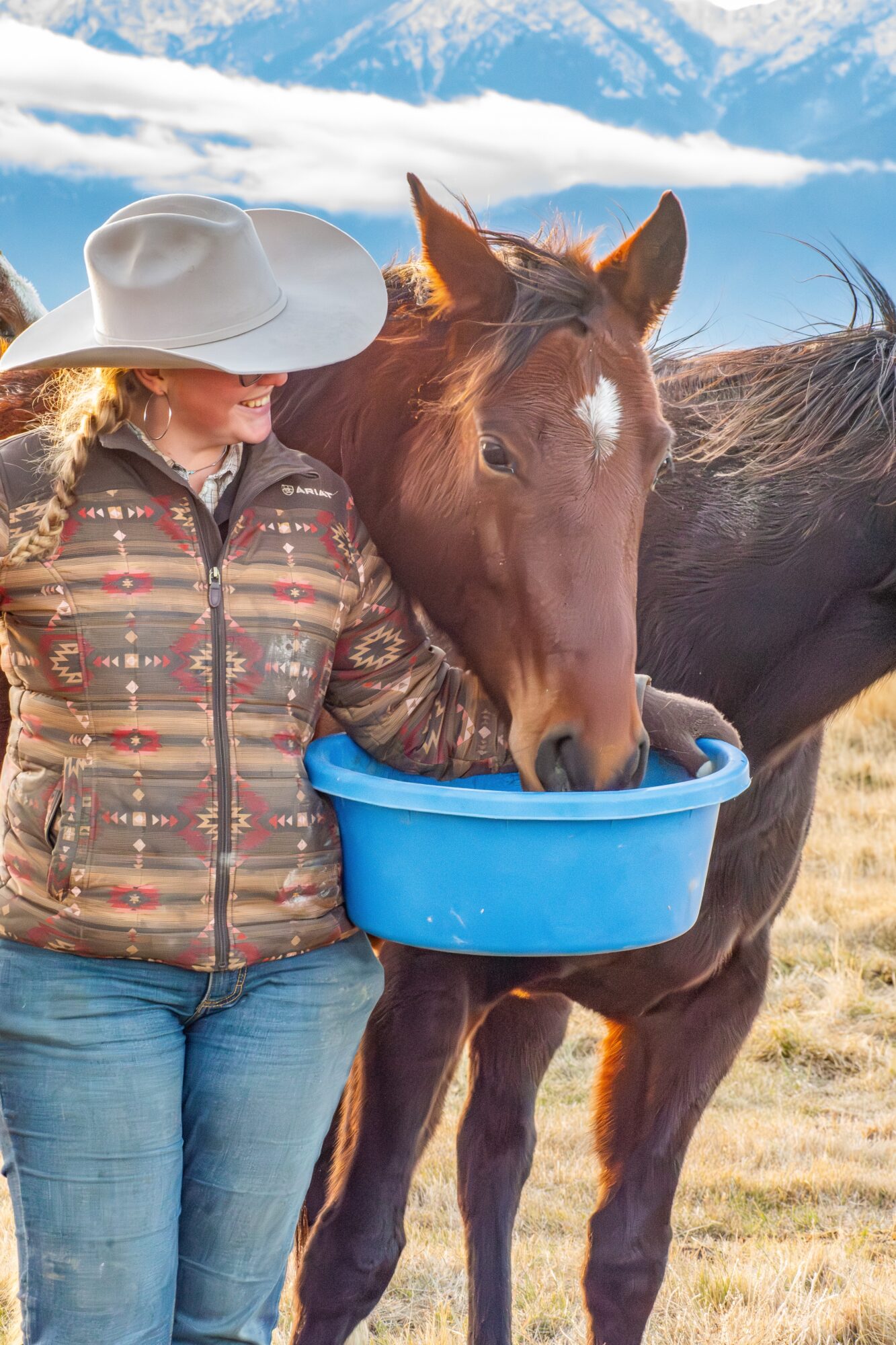 Person in a wide-brimmed hat feeds a horse from a blue bucket outdoors under a partly cloudy sky.