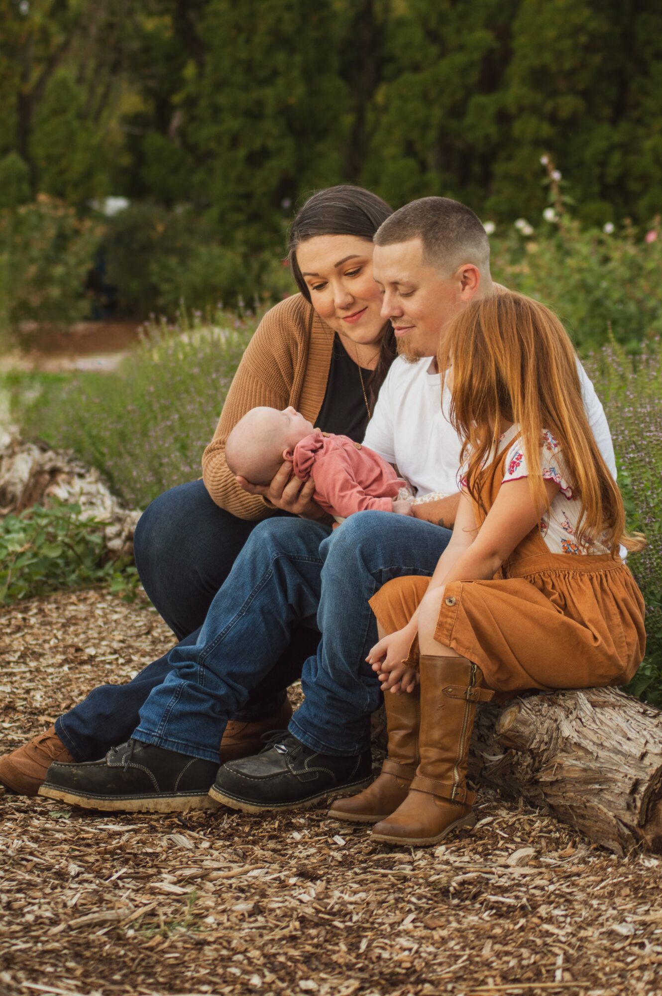 Family of four sitting outdoors on a log, with trees and greenery in the background, looking at a baby.