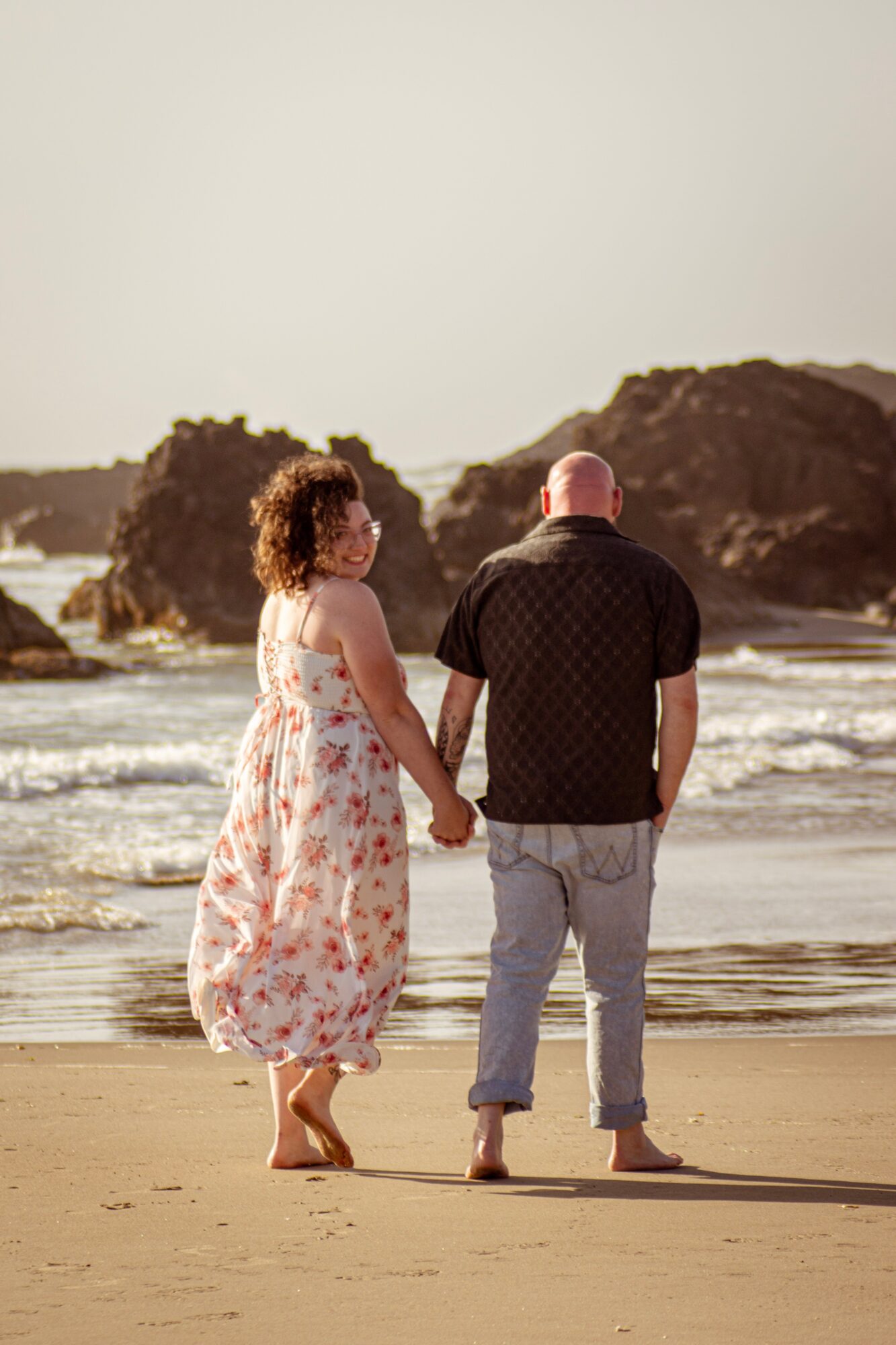 A couple holding hands walking on the beach with rocks and ocean in the background, woman in a floral dress, man in a black shirt and jeans.