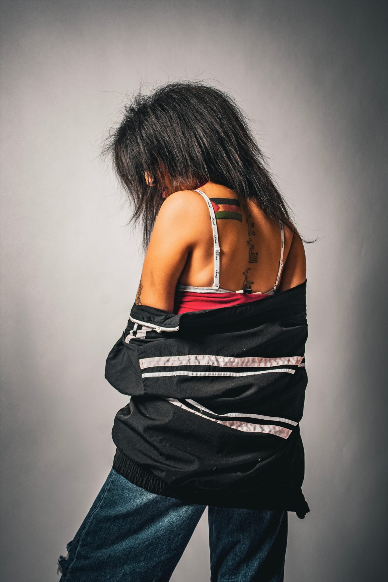 Person with dark, messy hair wearing a black jacket and jeans, facing away from the camera, against a plain background.