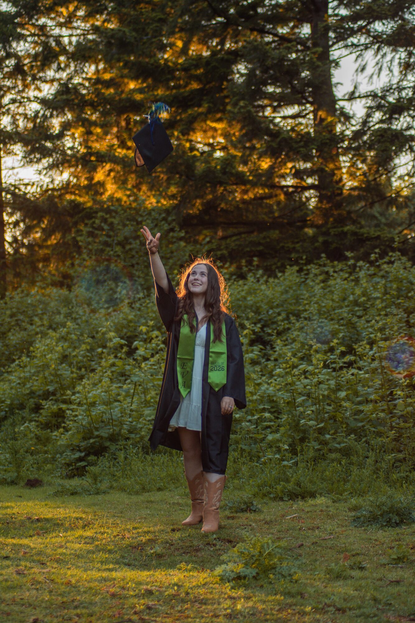 Person in graduation gown and cap outdoors, reaching up towards a flying cap, surrounded by trees and greenery.
