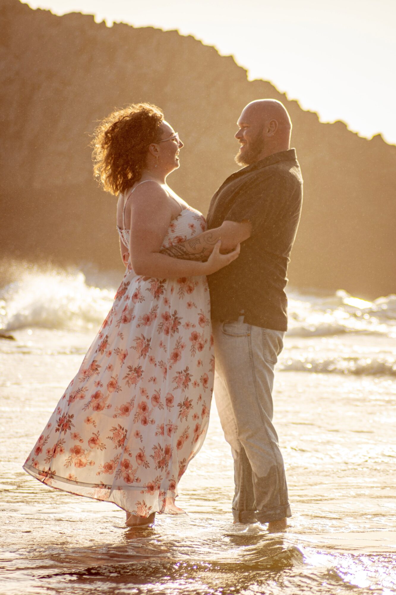 A couple stands in shallow water on a beach, holding hands and looking at each other, with a rocky hill in the background.