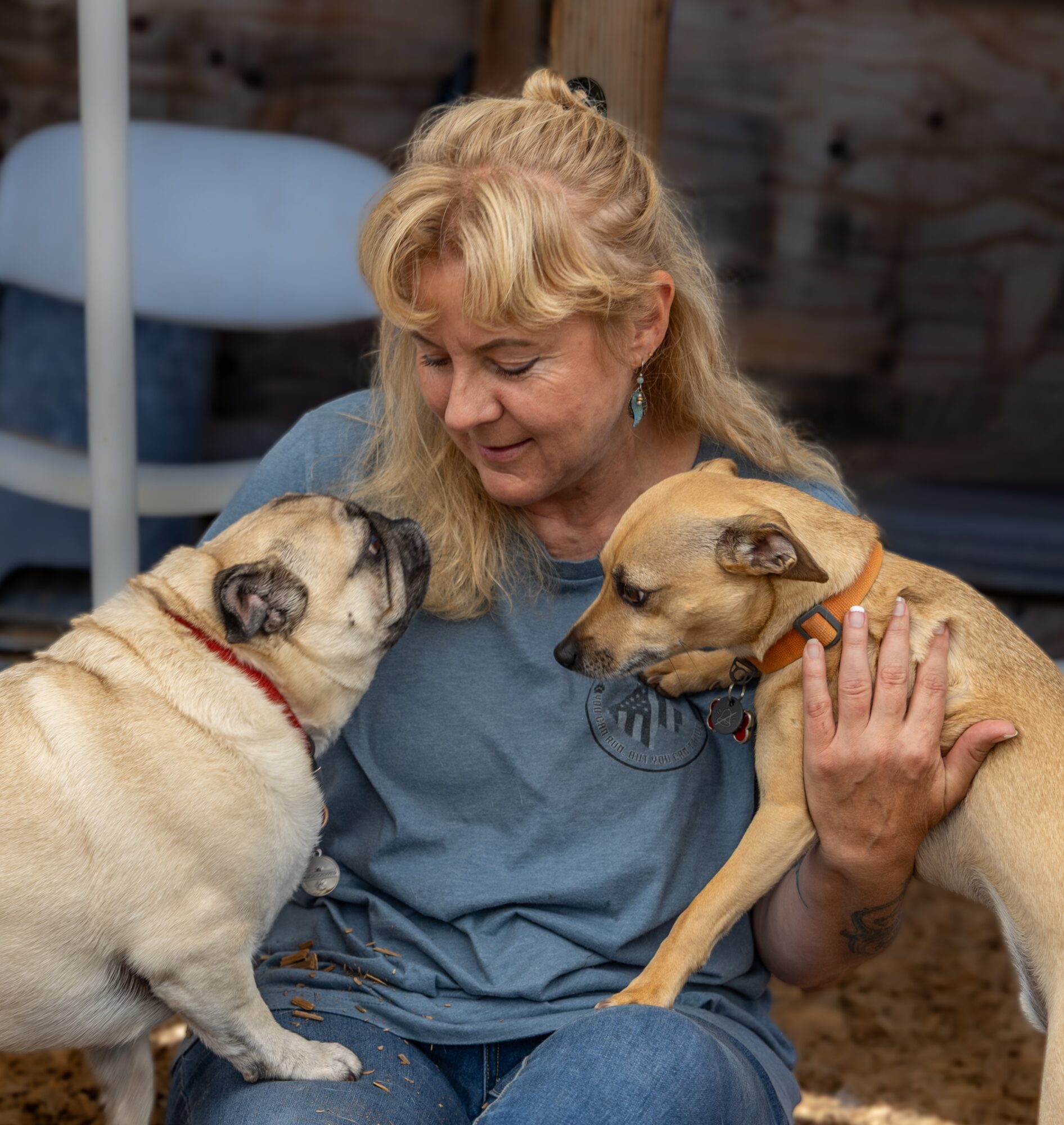 Woman holding two dogs close, one sniffing the other's face, in an outdoor setting with wooden background.