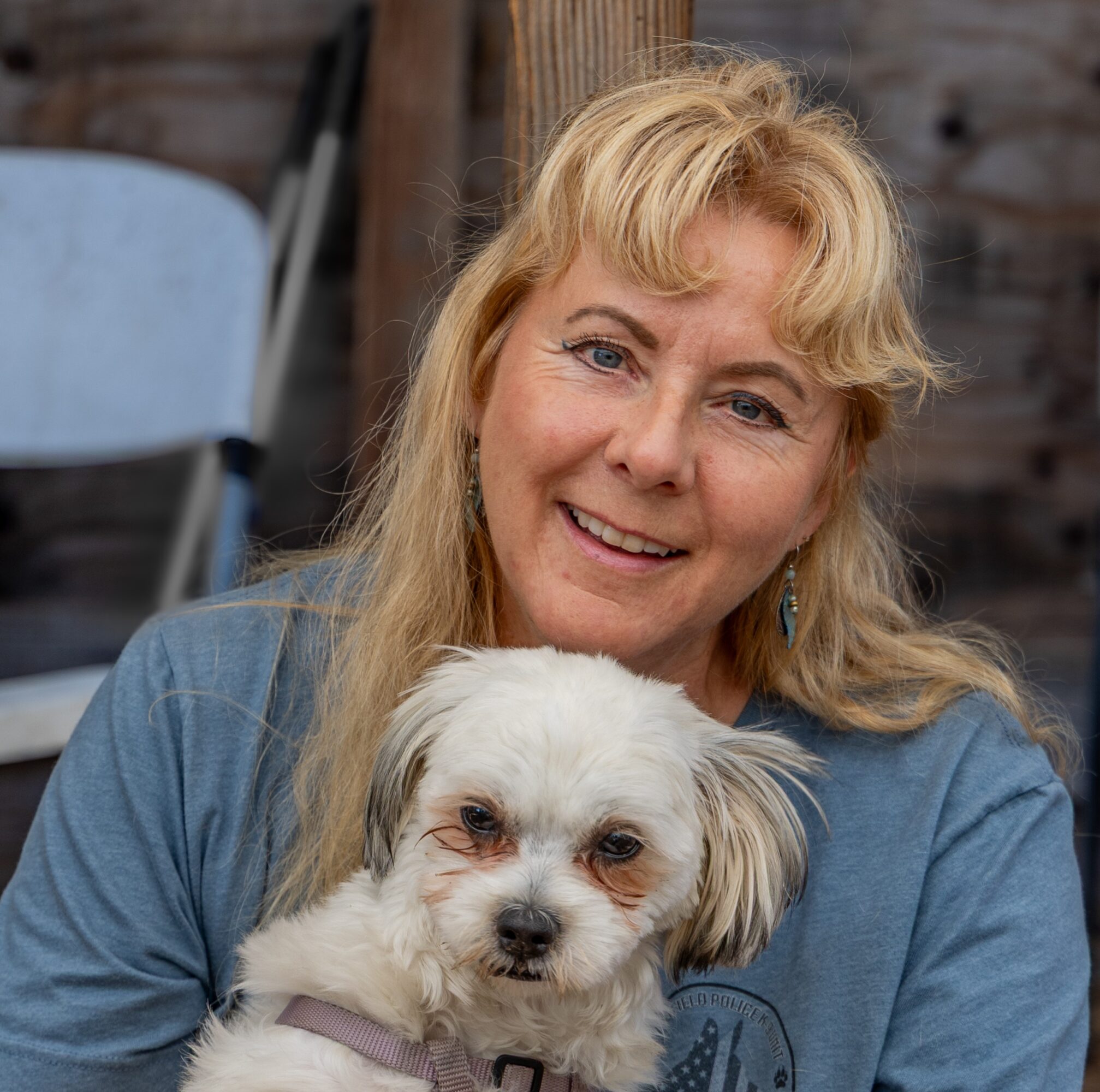 A woman with blonde hair smiling, holding a small white dog outdoors, wooden fence in background.