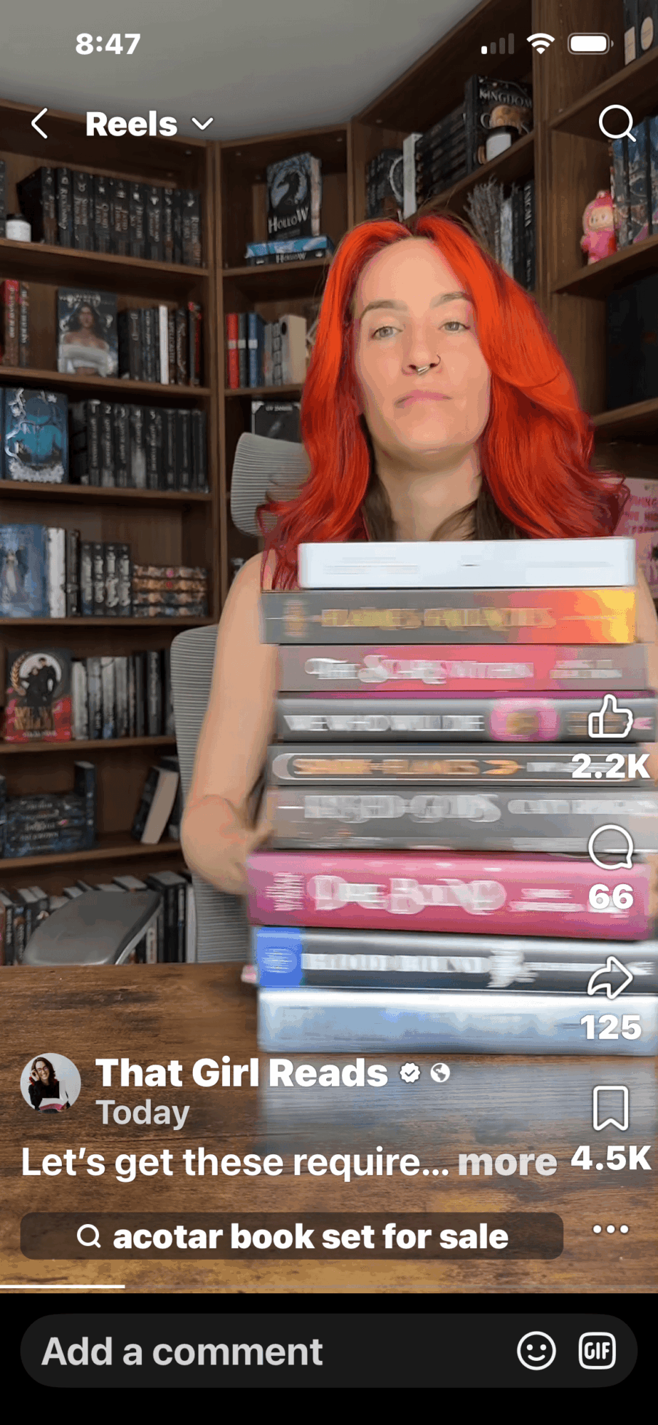 Woman with red hair holding a stack of books in a room with bookshelves behind her.