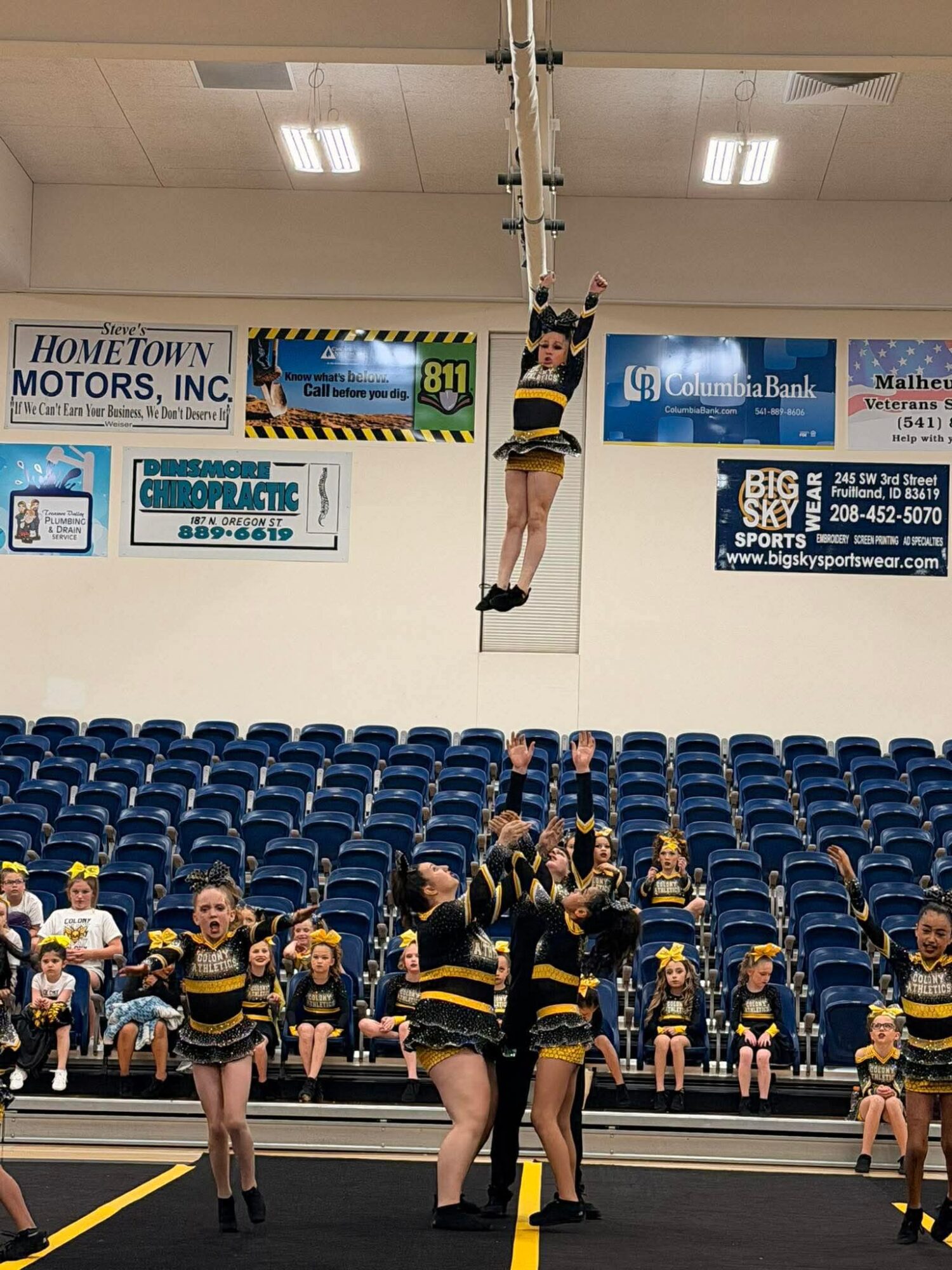 Cheerleader hanging from a rope above the gym floor, with other cheerleaders and spectators seated in the background.