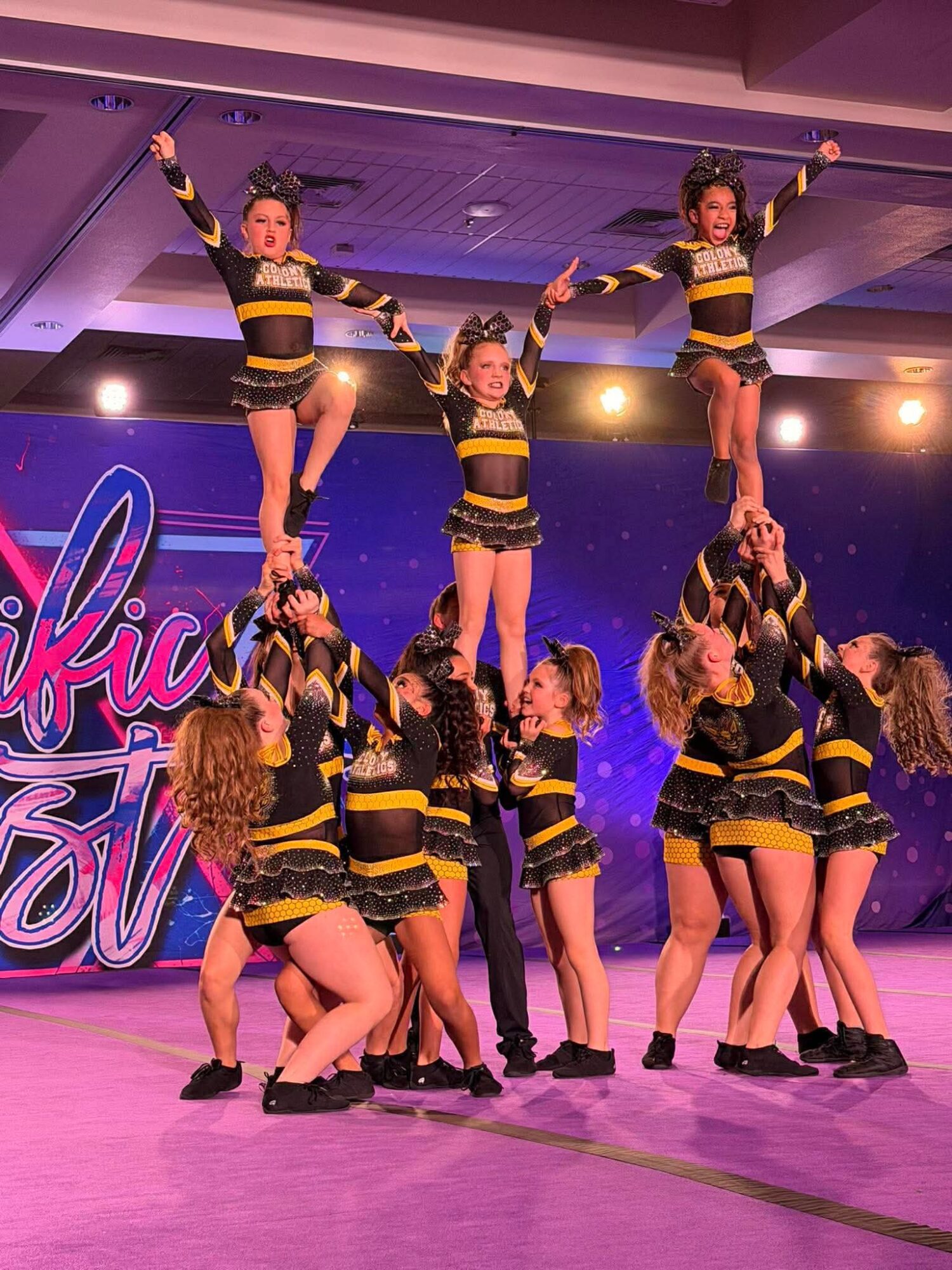 Group of young dancers performing a routine on stage, some lifted in the air, wearing matching black and yellow costumes.