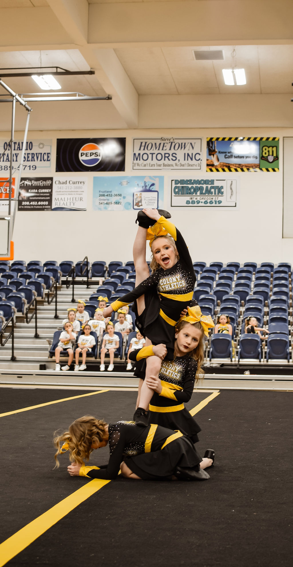 Two cheerleaders perform a stunt in a gymnasium with spectators seated in the background.