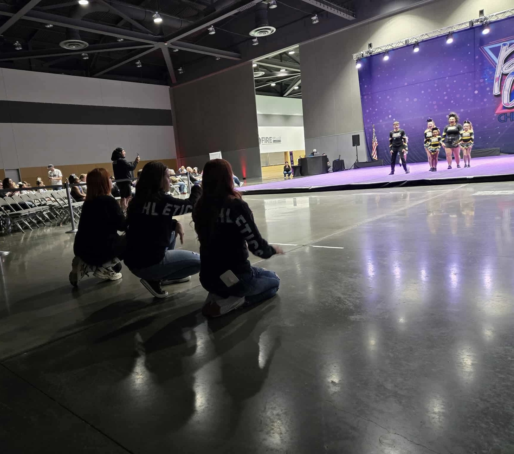 Three people kneeling on the floor watching a stage with performers, in a large indoor space with chairs and a purple backdrop.