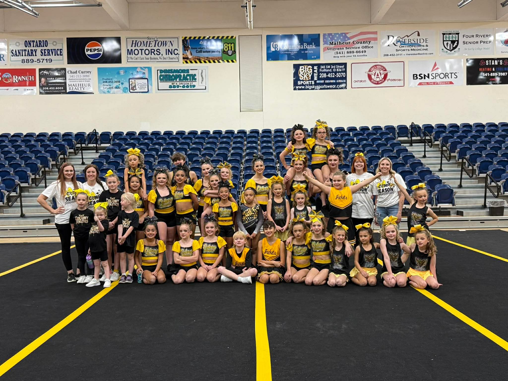Group of young cheerleaders in yellow and black uniforms posing on a gym floor with blue bleachers behind them.