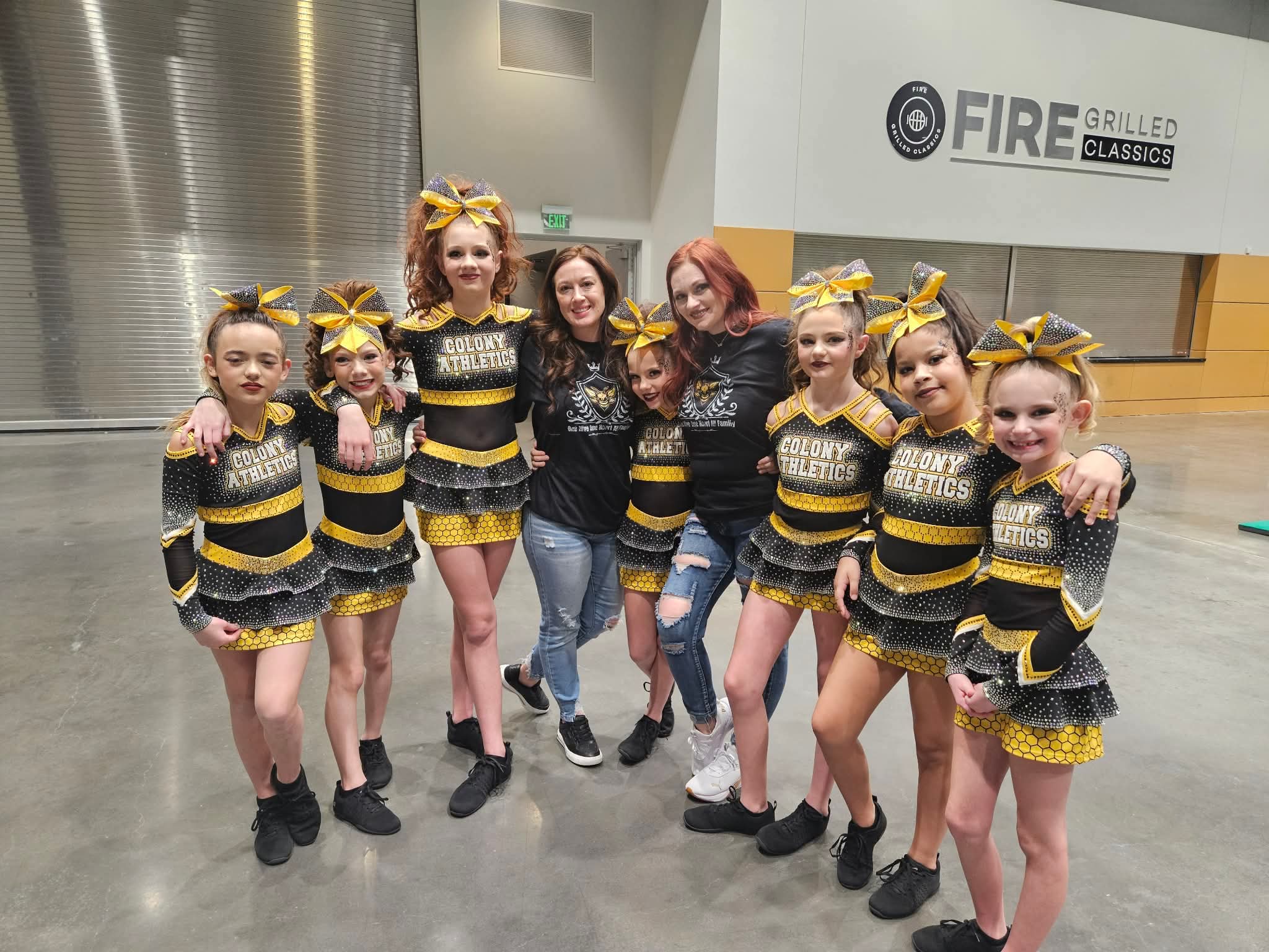 Group of young cheerleaders in matching black and yellow costumes with bows, posing with two adults indoors.