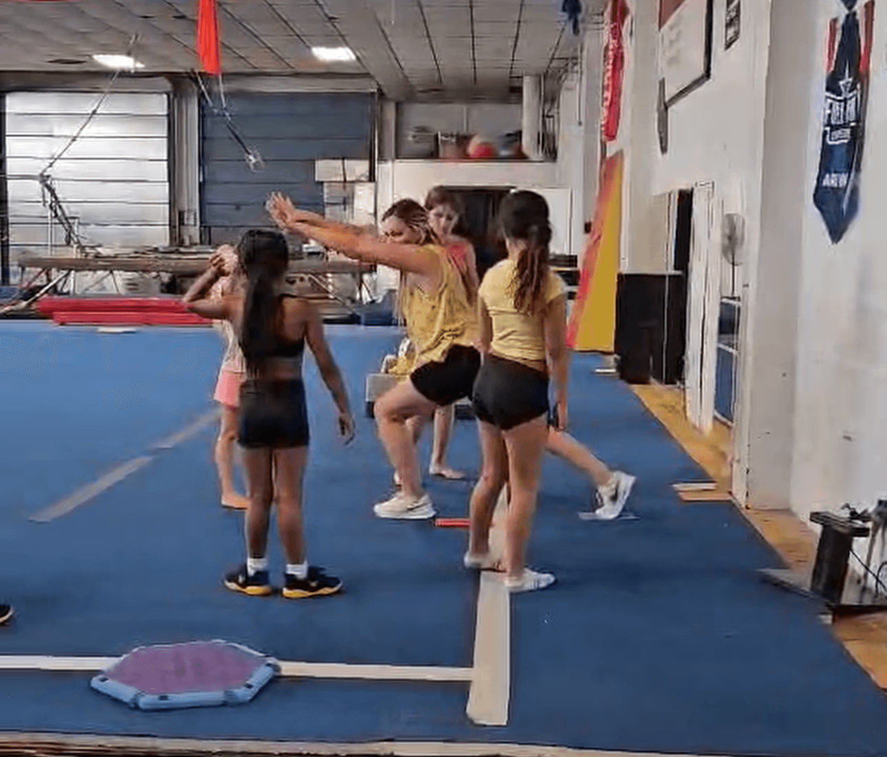 Group of young gymnasts practicing on a blue mat in a gym, with one person demonstrating a move.