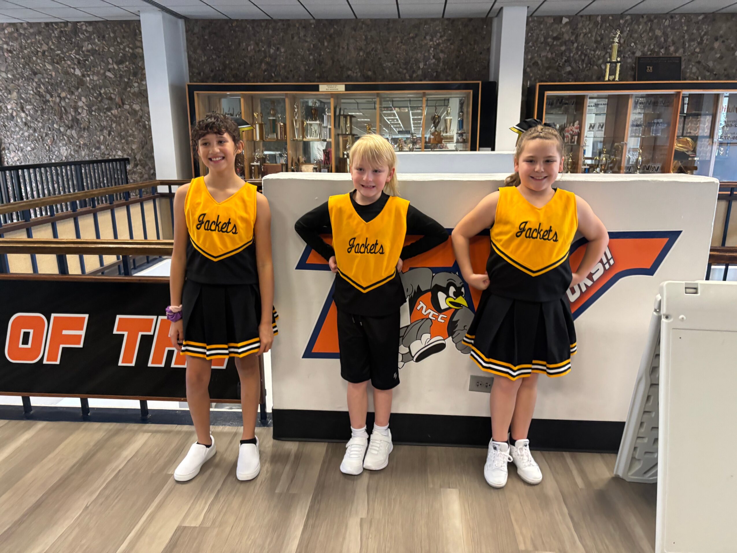 Three children in cheerleading uniforms standing indoors in front of a hockey arena entrance, smiling.
