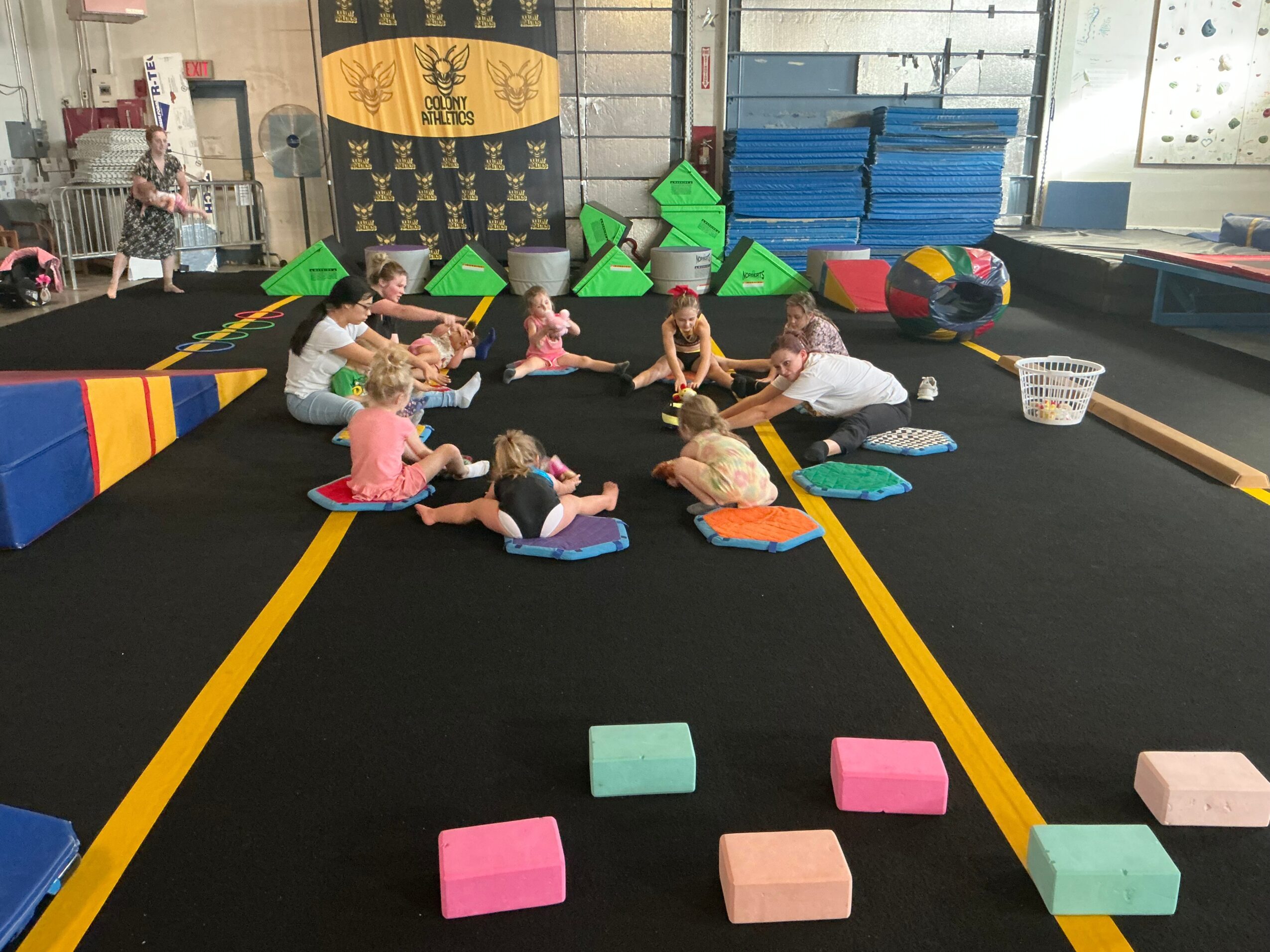 Children sit on mats in a gym, engaging in a group activity with colorful blocks and equipment around them.