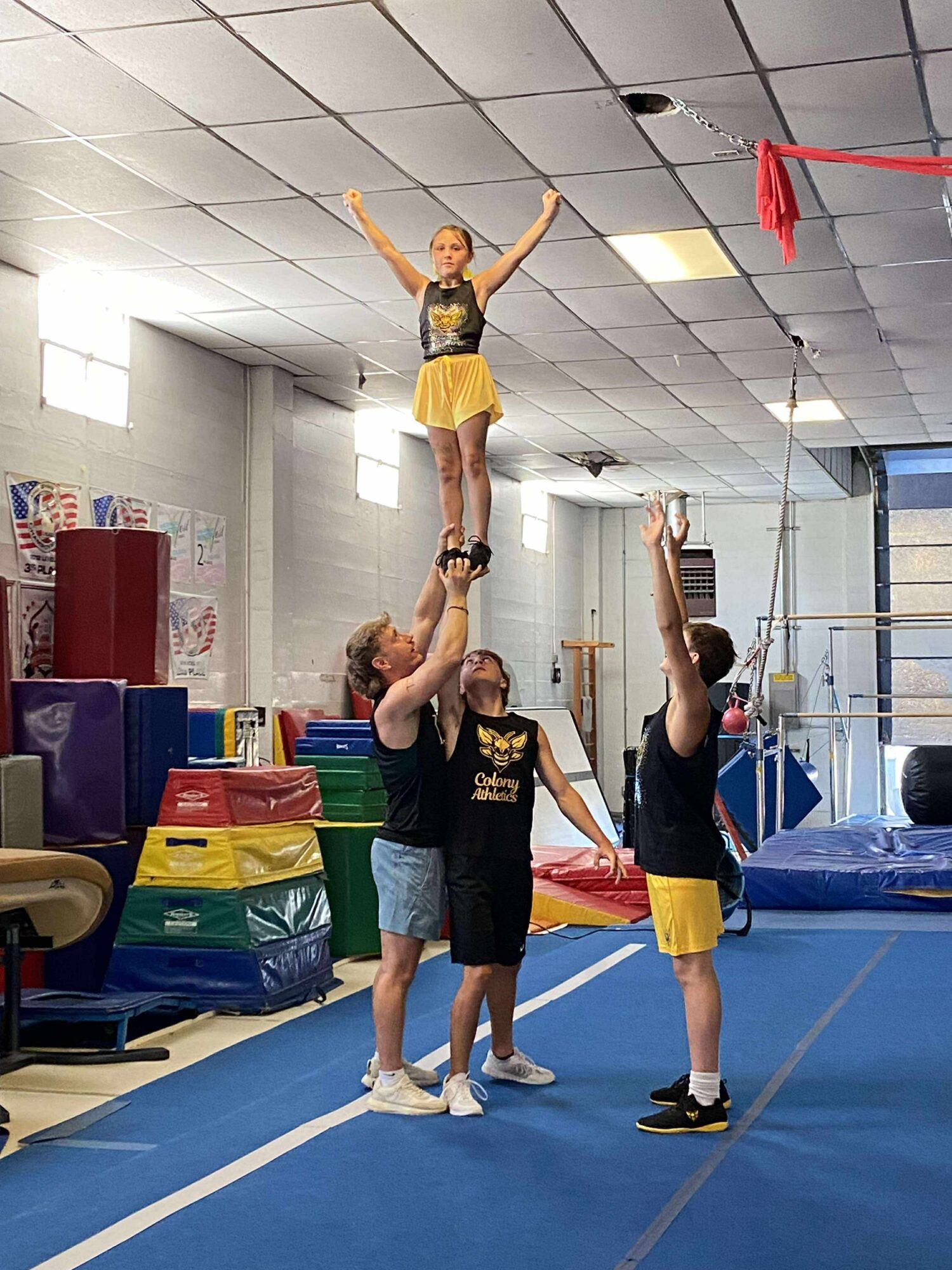 Three people in a gym, one person standing on the shoulders of two others, with arms raised, performing a cheerleading stunt.