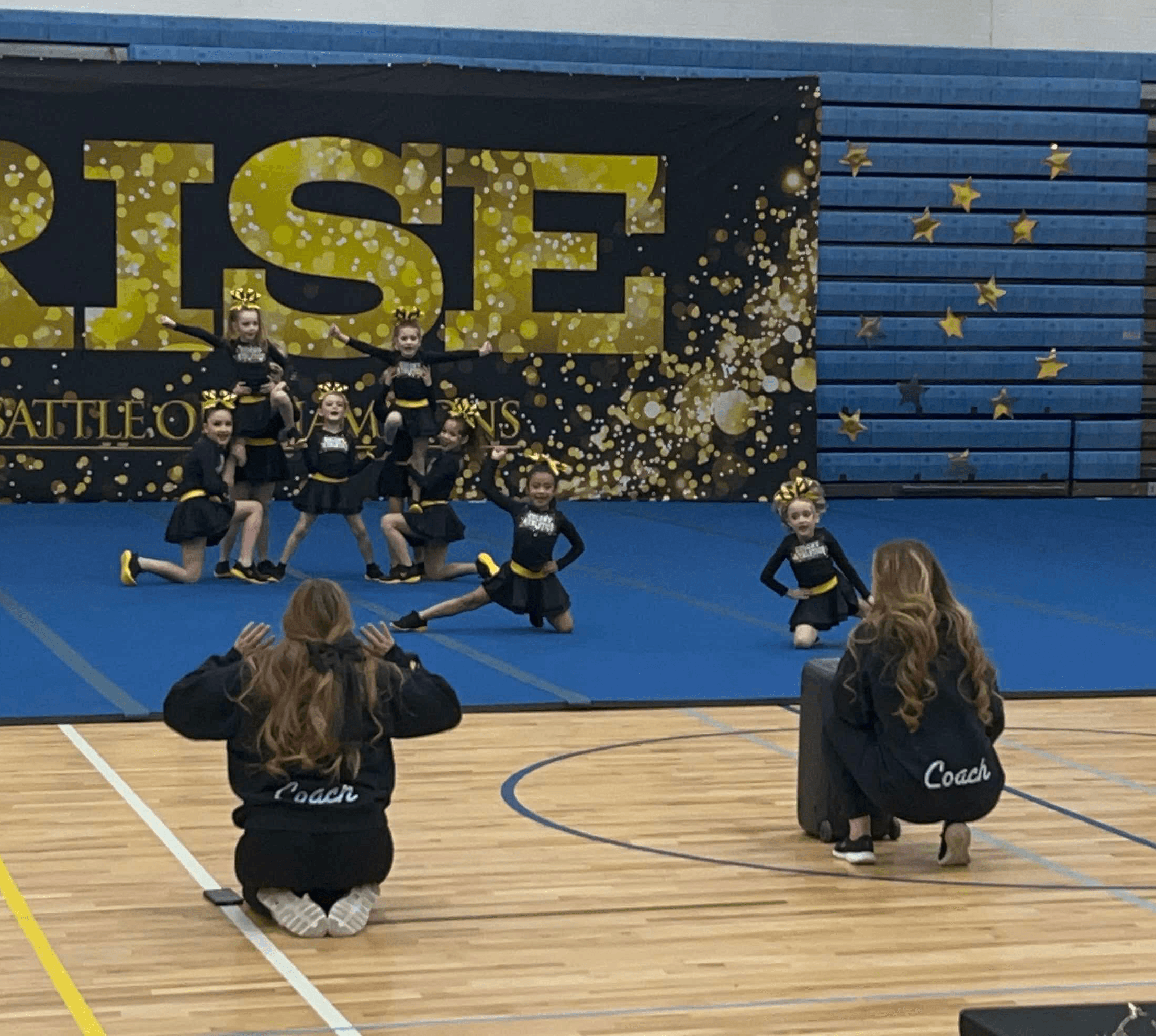 Group of young cheerleaders performing stunts on a blue mat, with two coaches kneeling in front, in a gymnasium.