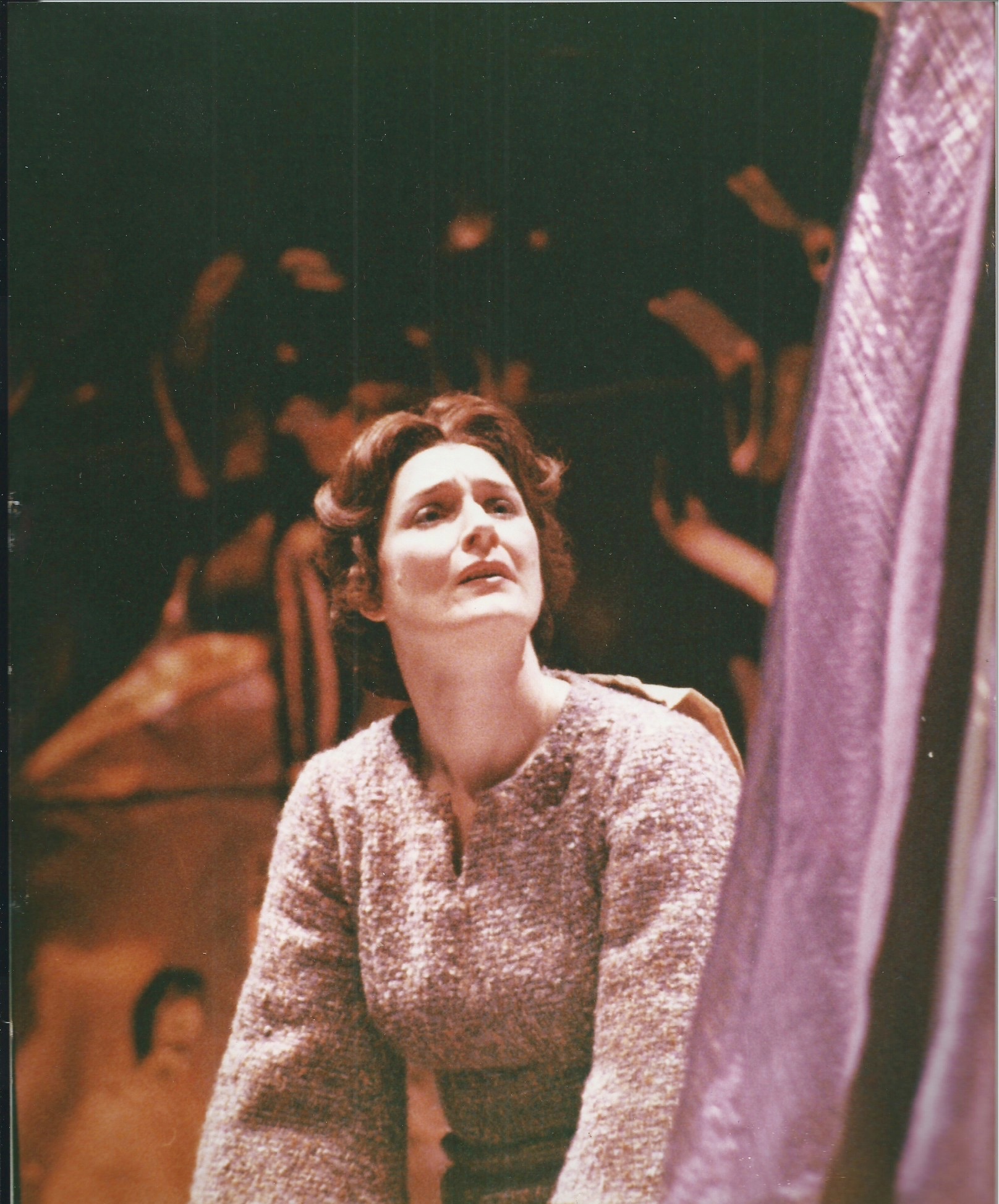 Woman with dark hair and light-colored sweater looking upward, with a curtain partially visible on the right side.