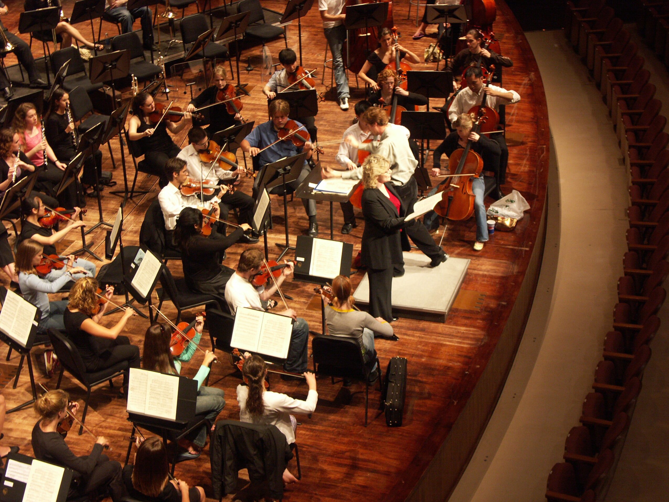 Orchestra performing on stage with musicians playing string instruments, conductor in front, wooden stage, audience seating visible on right.