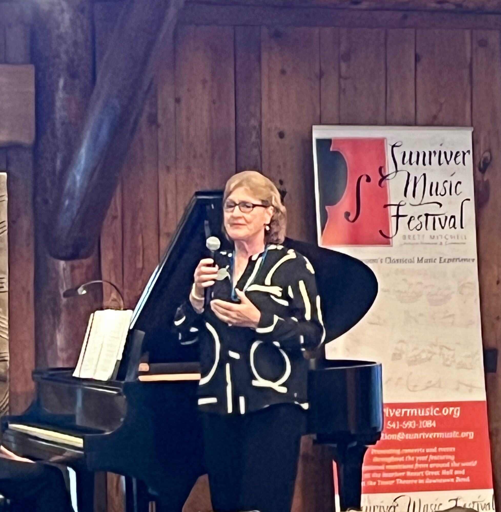 Woman speaking into microphone at piano, wooden wall background, Sunriver Music Festival poster nearby.