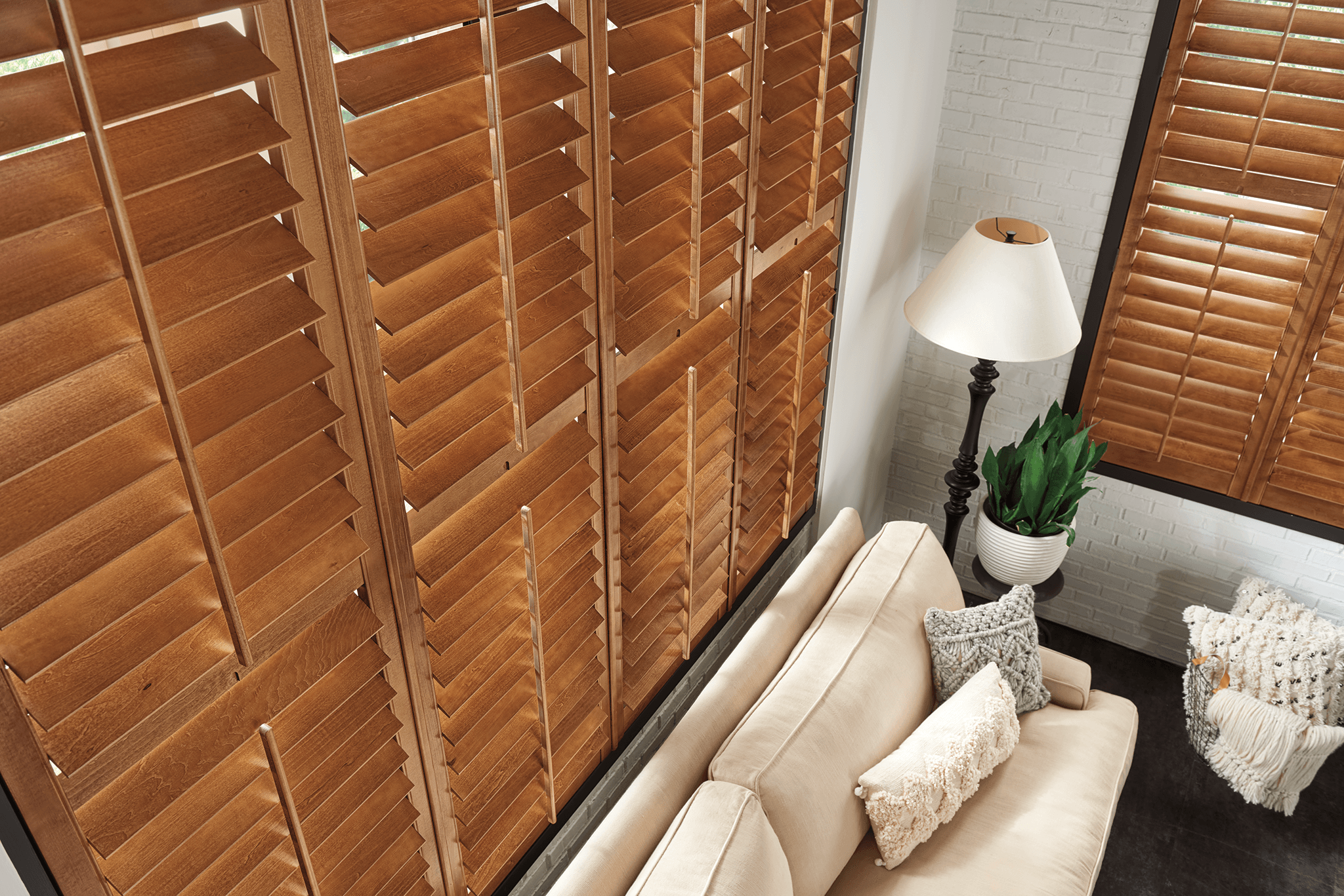 Upright view of wooden window shutters, a beige sofa with pillows, a white lamp, and a potted plant in a cozy room.
