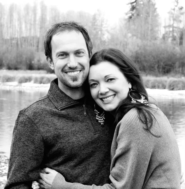 A smiling man and woman pose outdoors, close together, with trees and a body of water in the background.