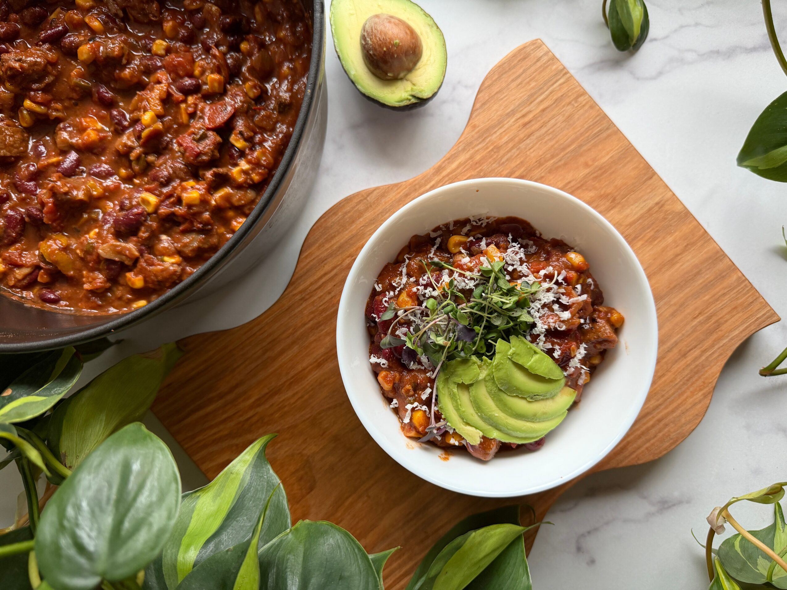 Bowl of chili with avocado slices and cilantro on a wooden board, surrounded by avocado halves and green leaves.