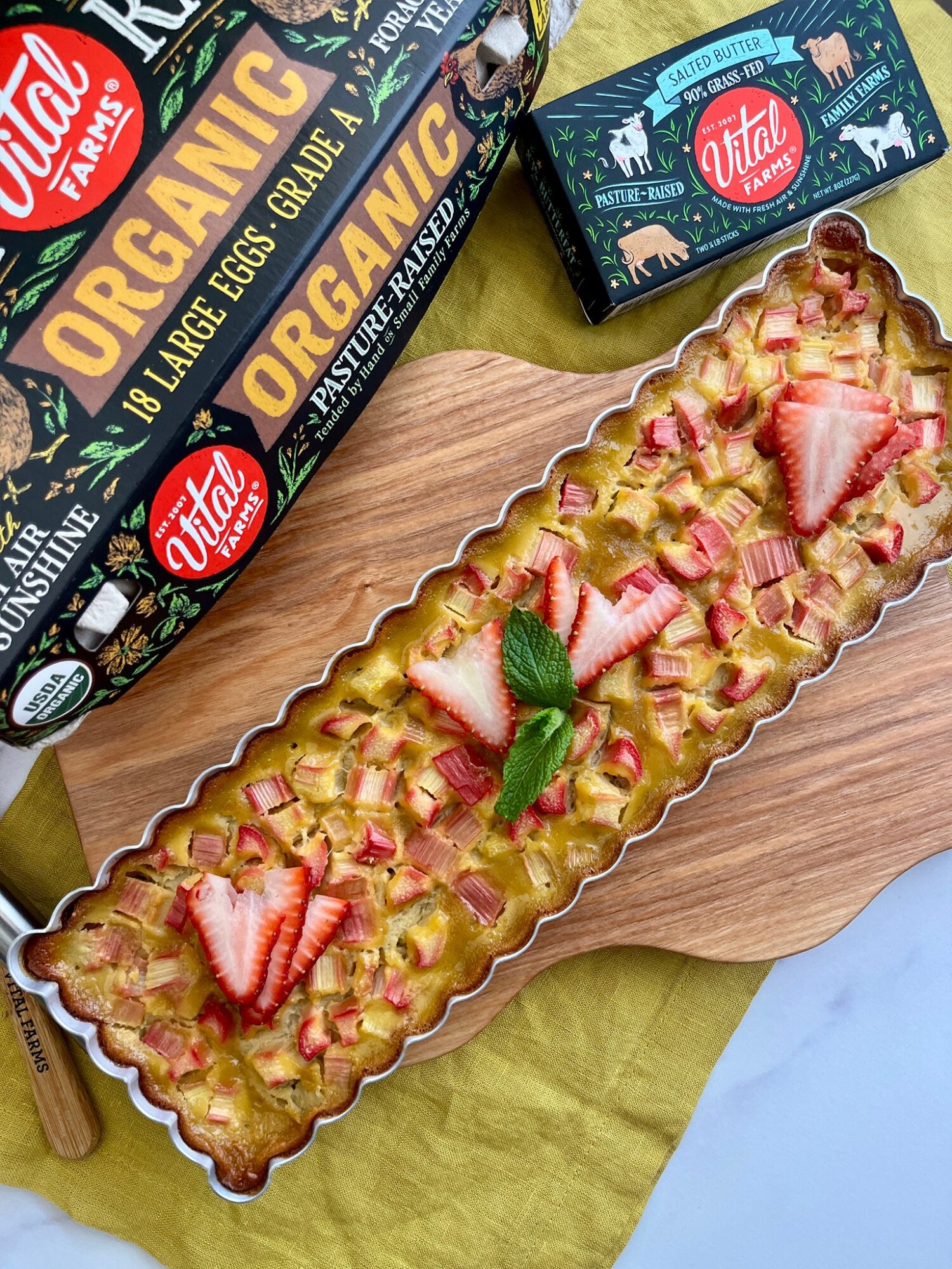 Long rectangular tart topped with sliced strawberries, mint leaves, and a lattice crust, on a wooden board with boxes of eggs nearby.